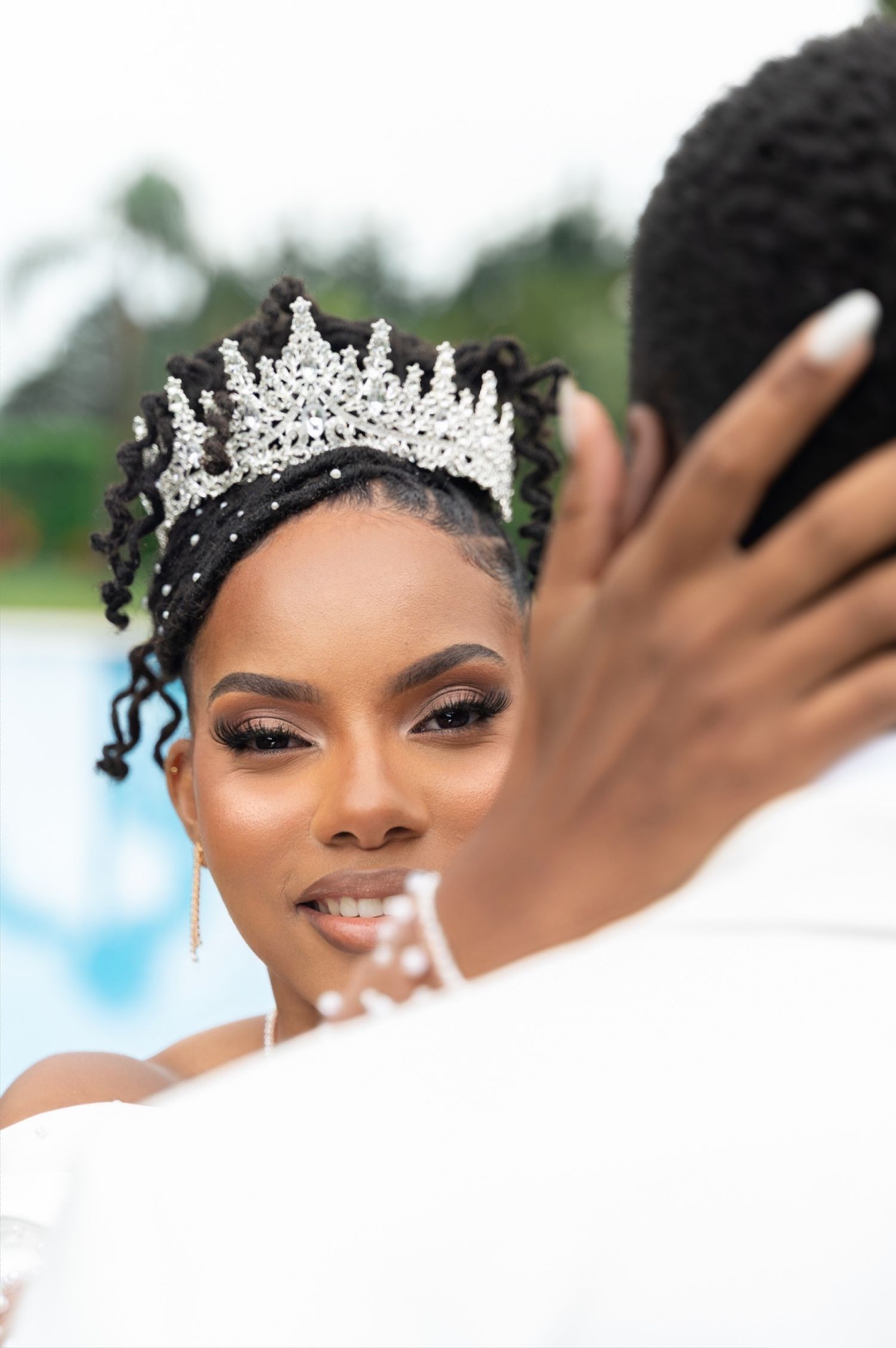 Close-up of radiant Black bride in diamond crown smiling over groom's shoulder at outdoor wedding.
