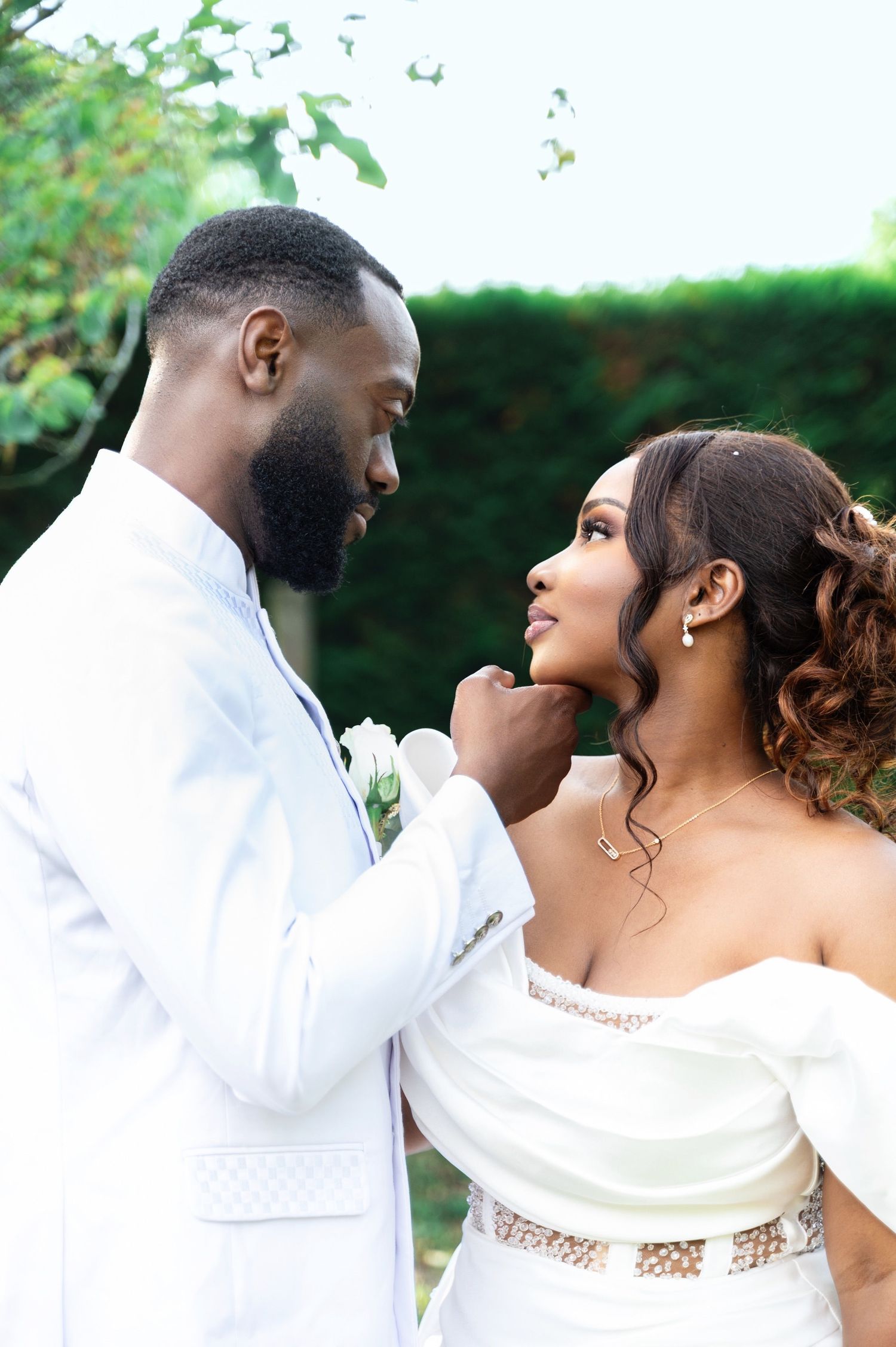 A Black groom in a white suit gently lifts his bride's chin as they share an intimate moment outdoors.