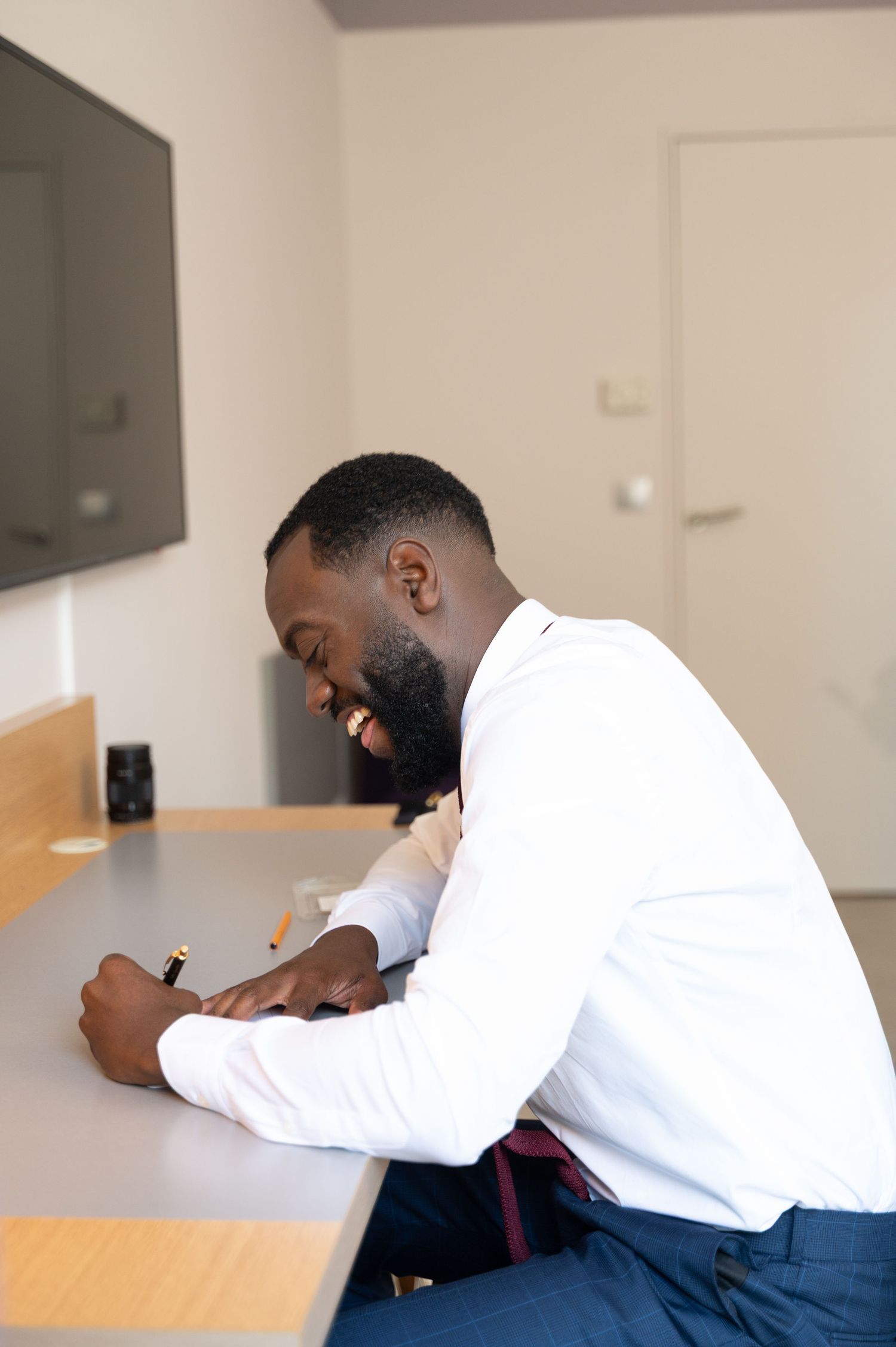 Smiling Black man in white dress shirt laughing while writing at a desk in a bright modern room.