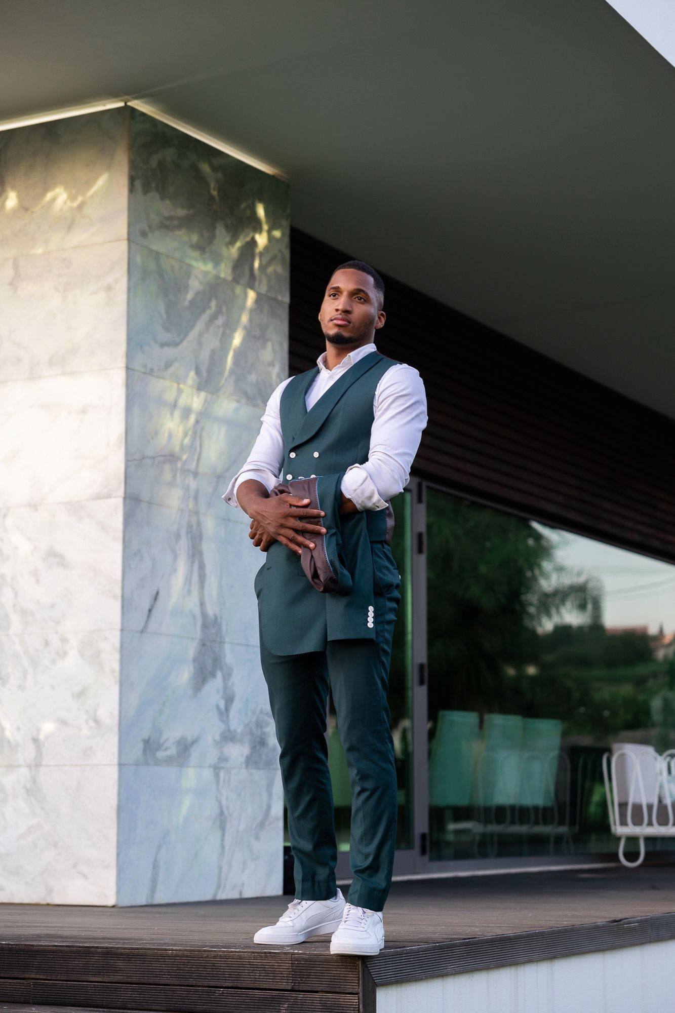 Stylish Black man in green vest suit holding phone standing outside modern building.