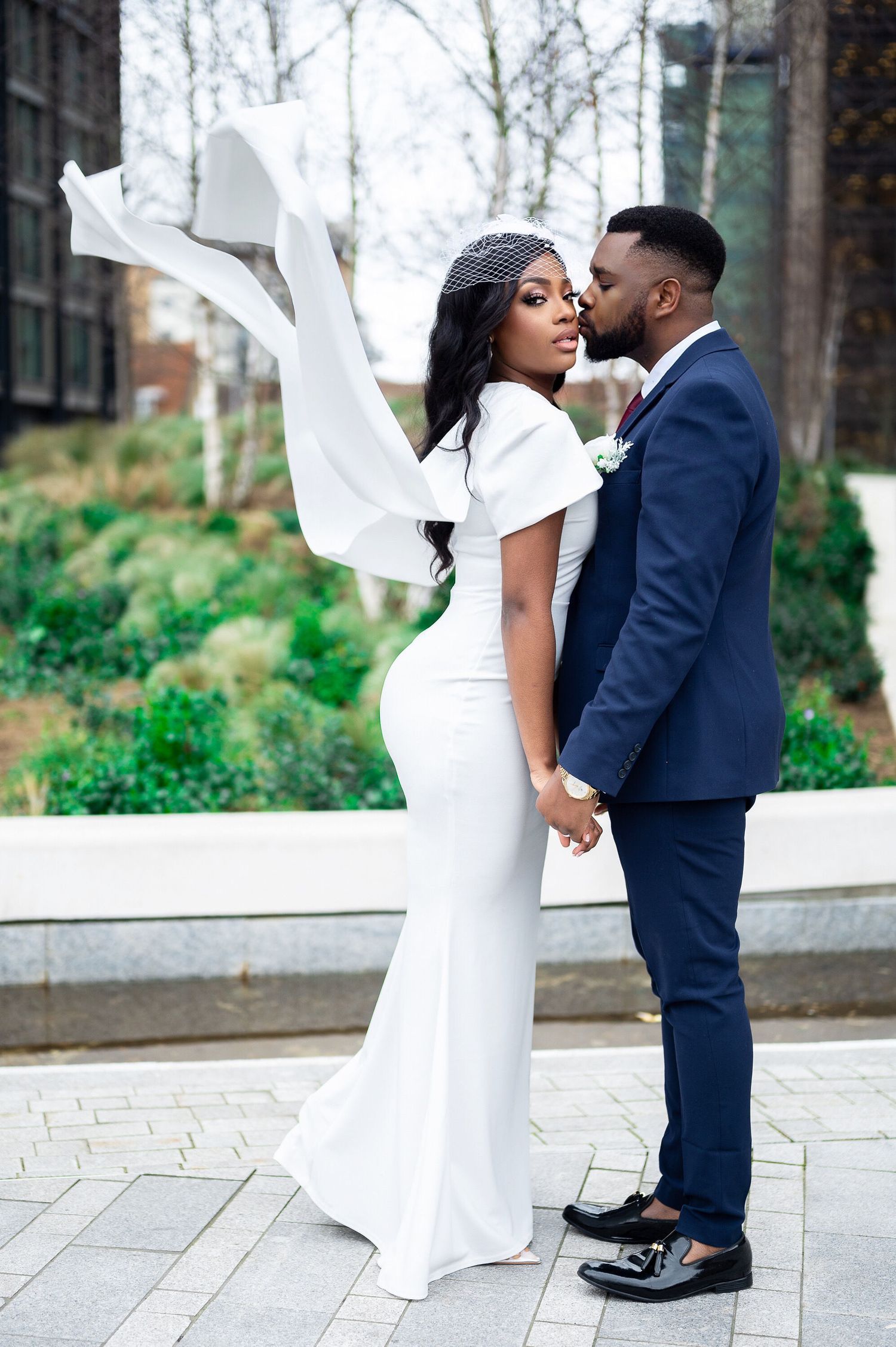 A Black bride and groom pose romantically outdoors, her white gown and veil flowing in the breeze.