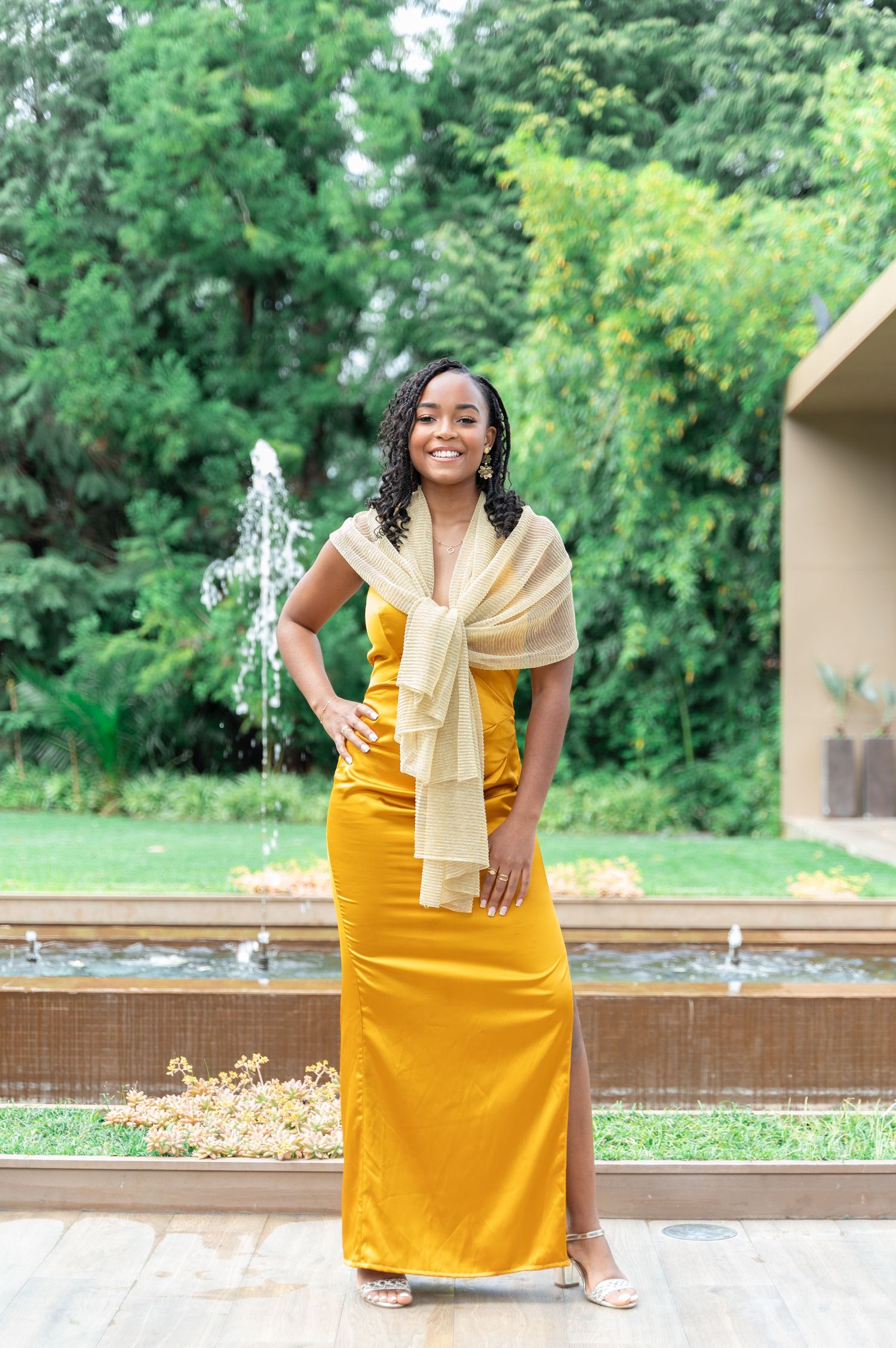 Elegant woman in mustard yellow maxi dress with gold wrap posing by fountain.