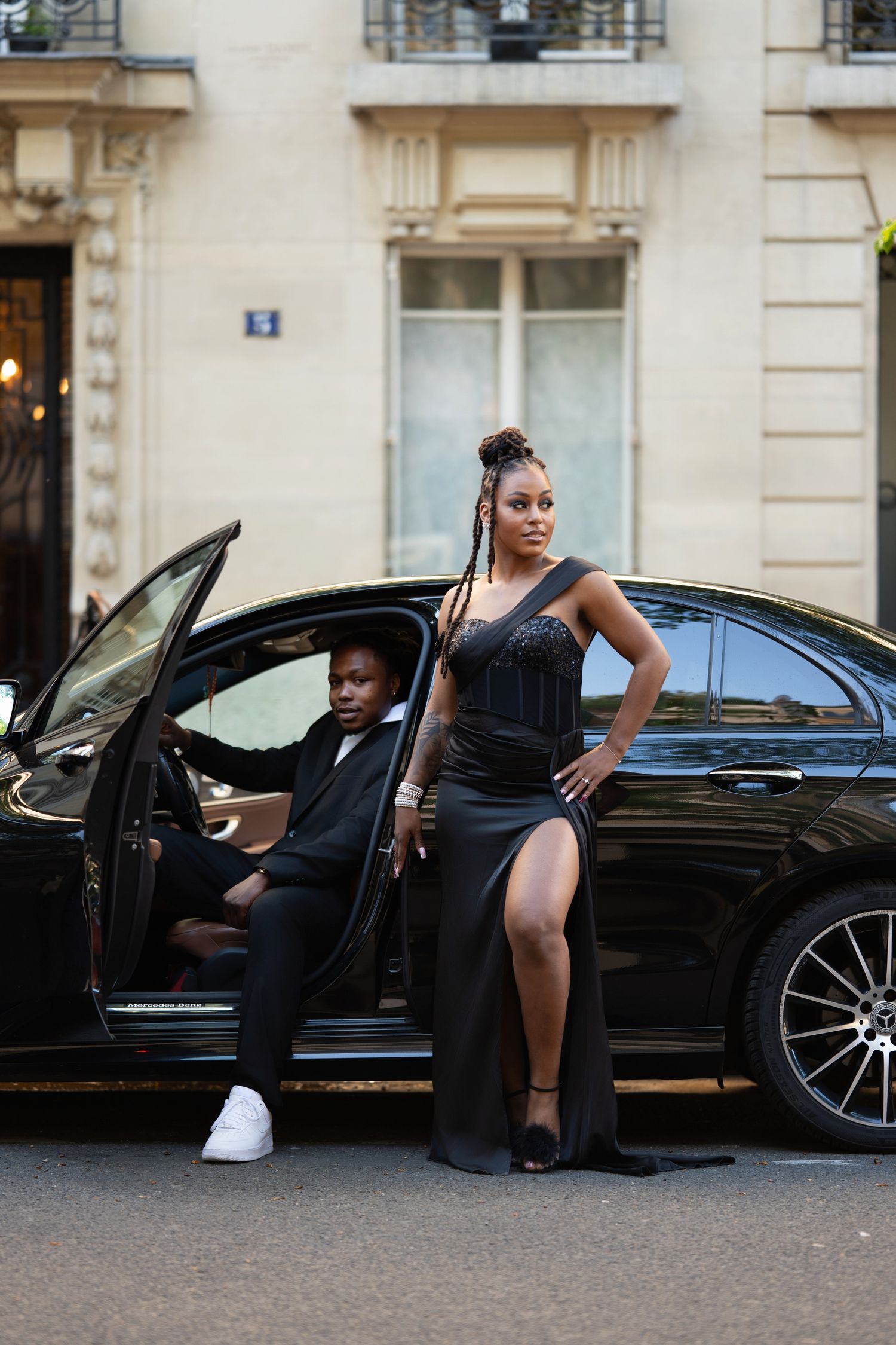 Elegant couple in black formal attire posing beside a luxury black car on a Parisian street.