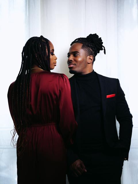 Couple in coordinating burgundy and black outfits gazing at each other against white curtain backdrop.