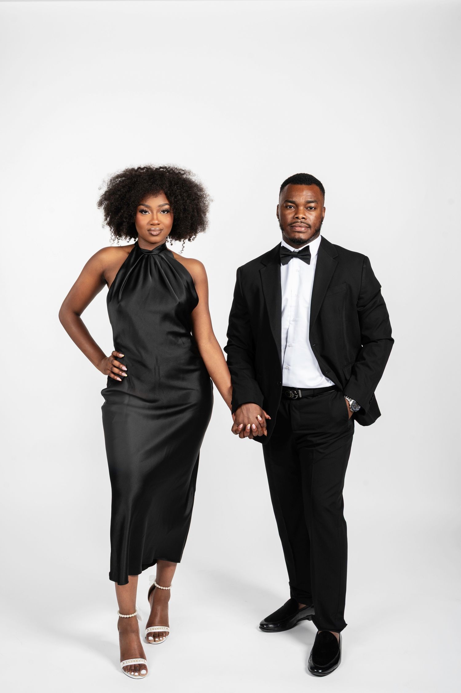 An elegant Black couple in matching black formal attire hold hands while posing together against a white background.