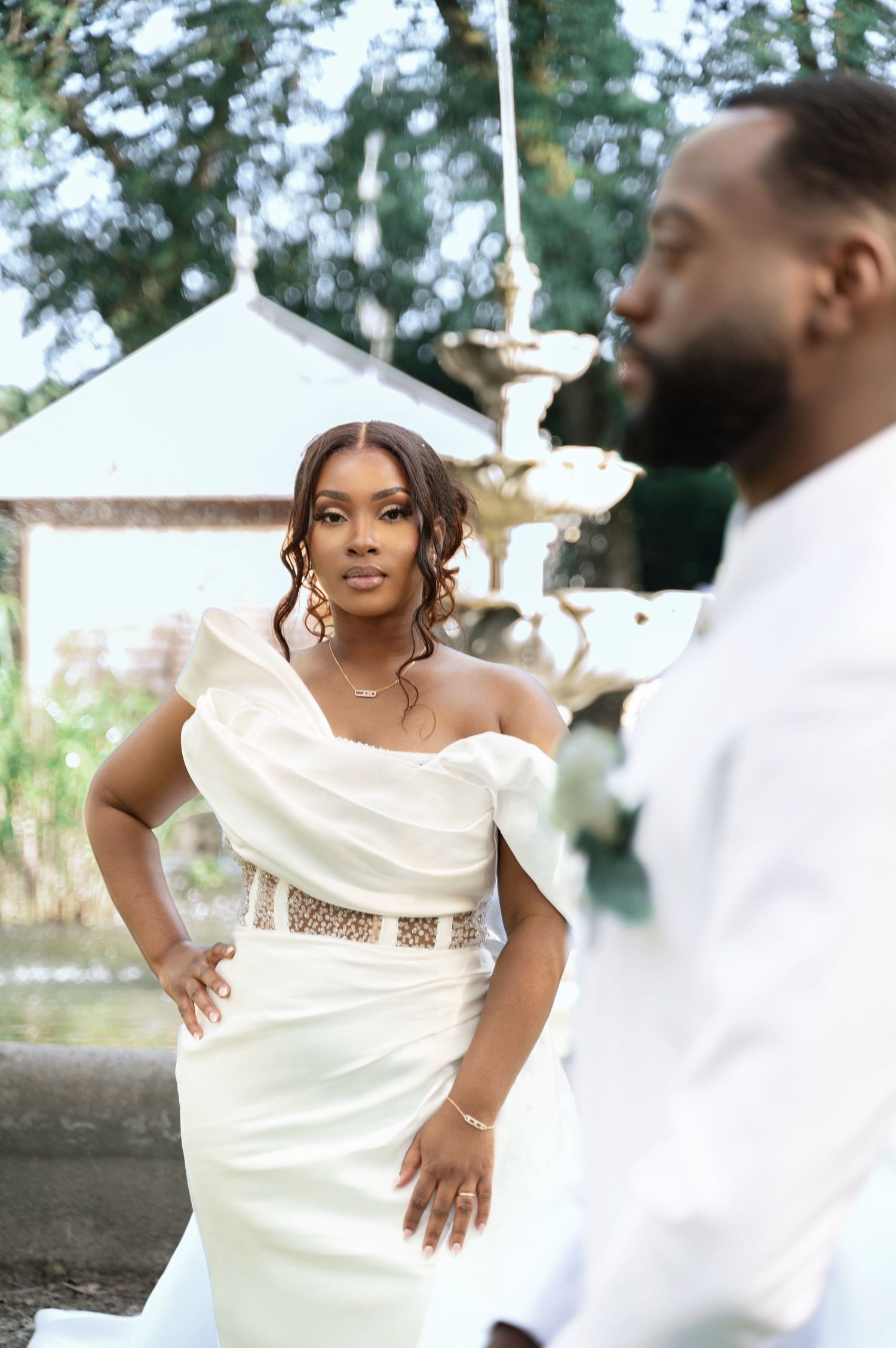 Beautiful bride in off-shoulder white gown with gold belt posing near a stone fountain at wedding venue.