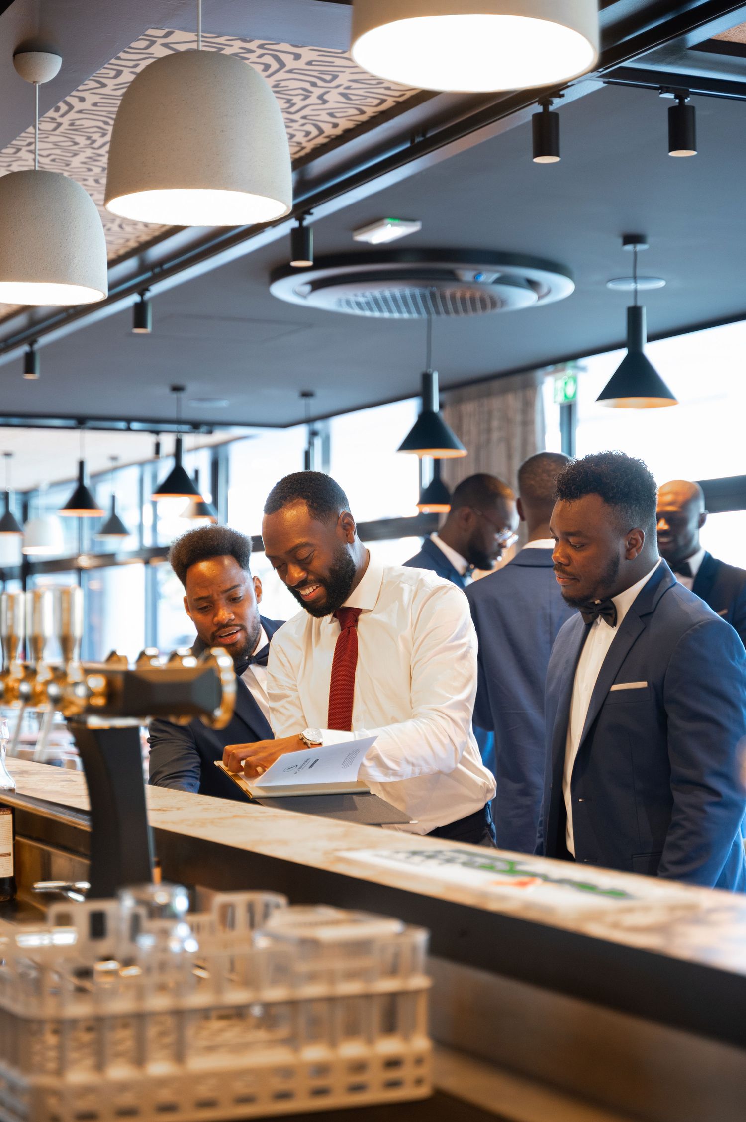 Group of well-dressed Black men in suits gather and smile around a modern restaurant bar.