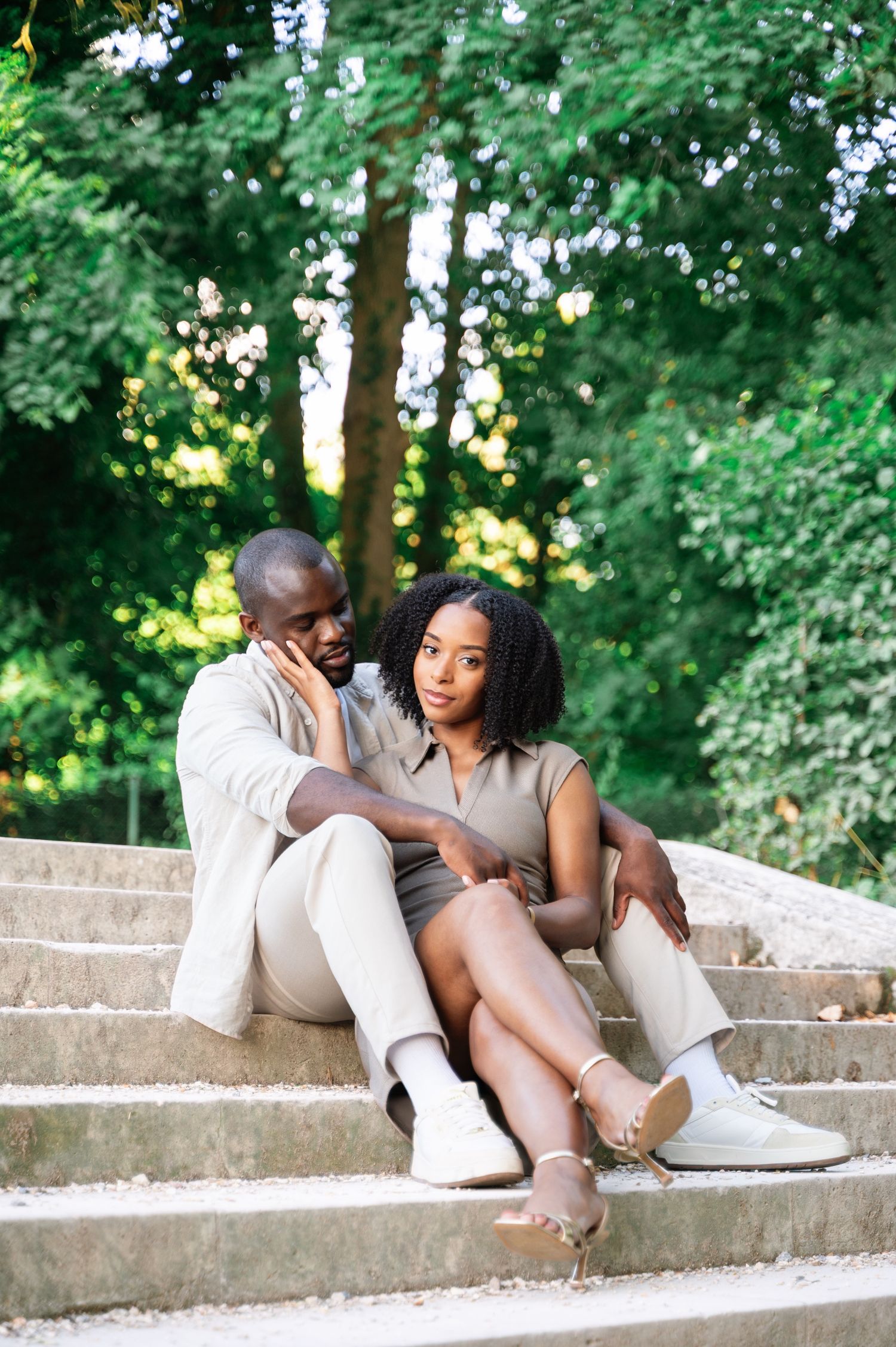 A Black couple sits together on stone steps in a green park, sharing a quiet and intimate moment.