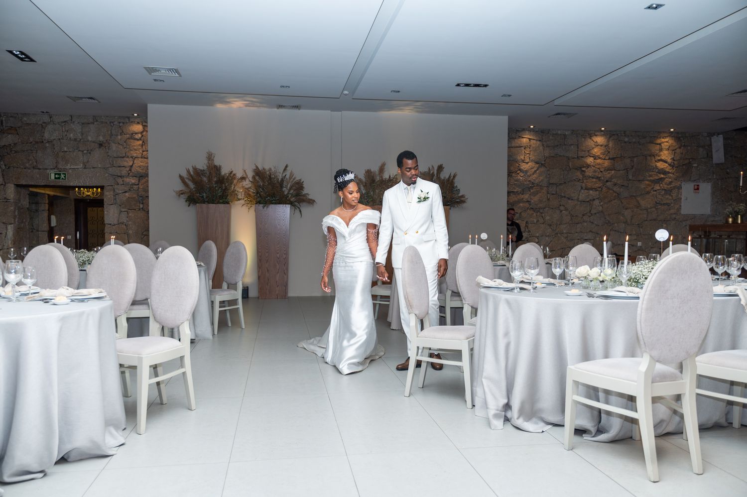 Newlyweds walk through a sophisticated white stone wedding venue with elegant table settings and floral arrangements.