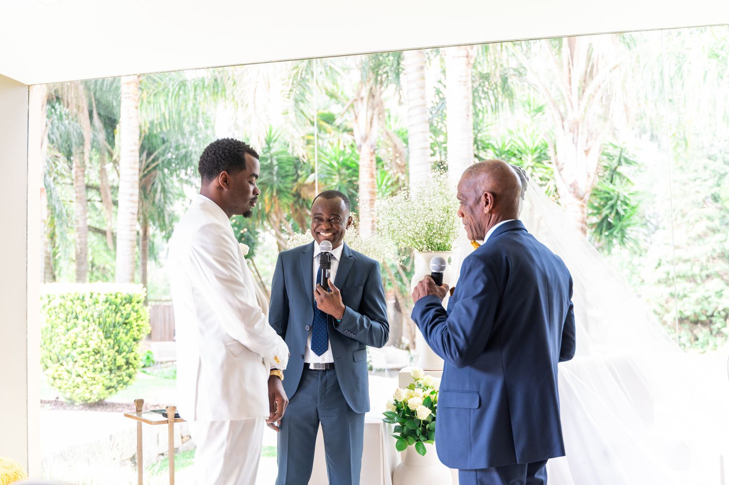 Three men in suits converse during a wedding ceremony in a bright venue with tropical greenery.