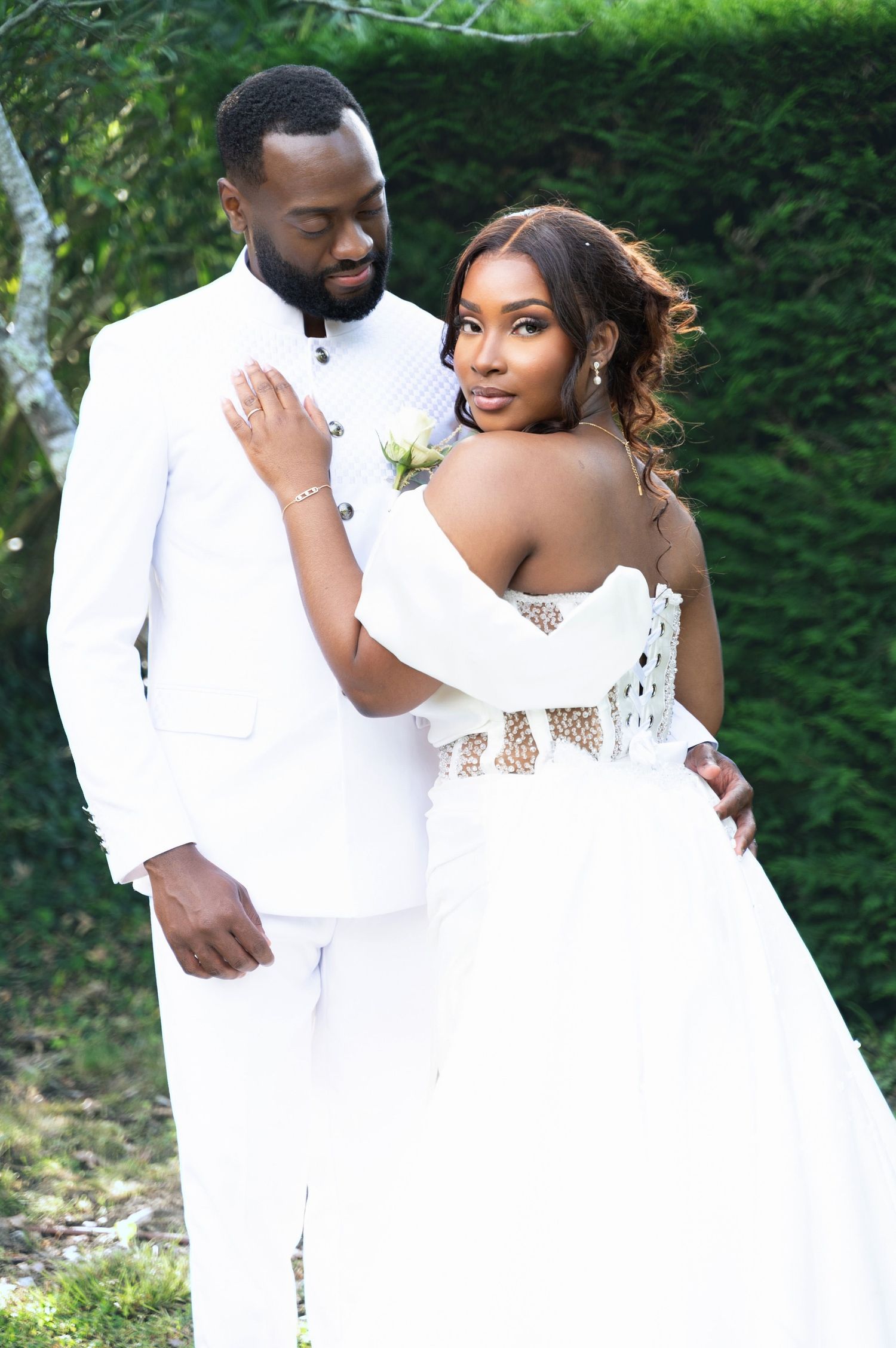 A Black bride in a white off-shoulder gown leans against her groom while posing for wedding photos.