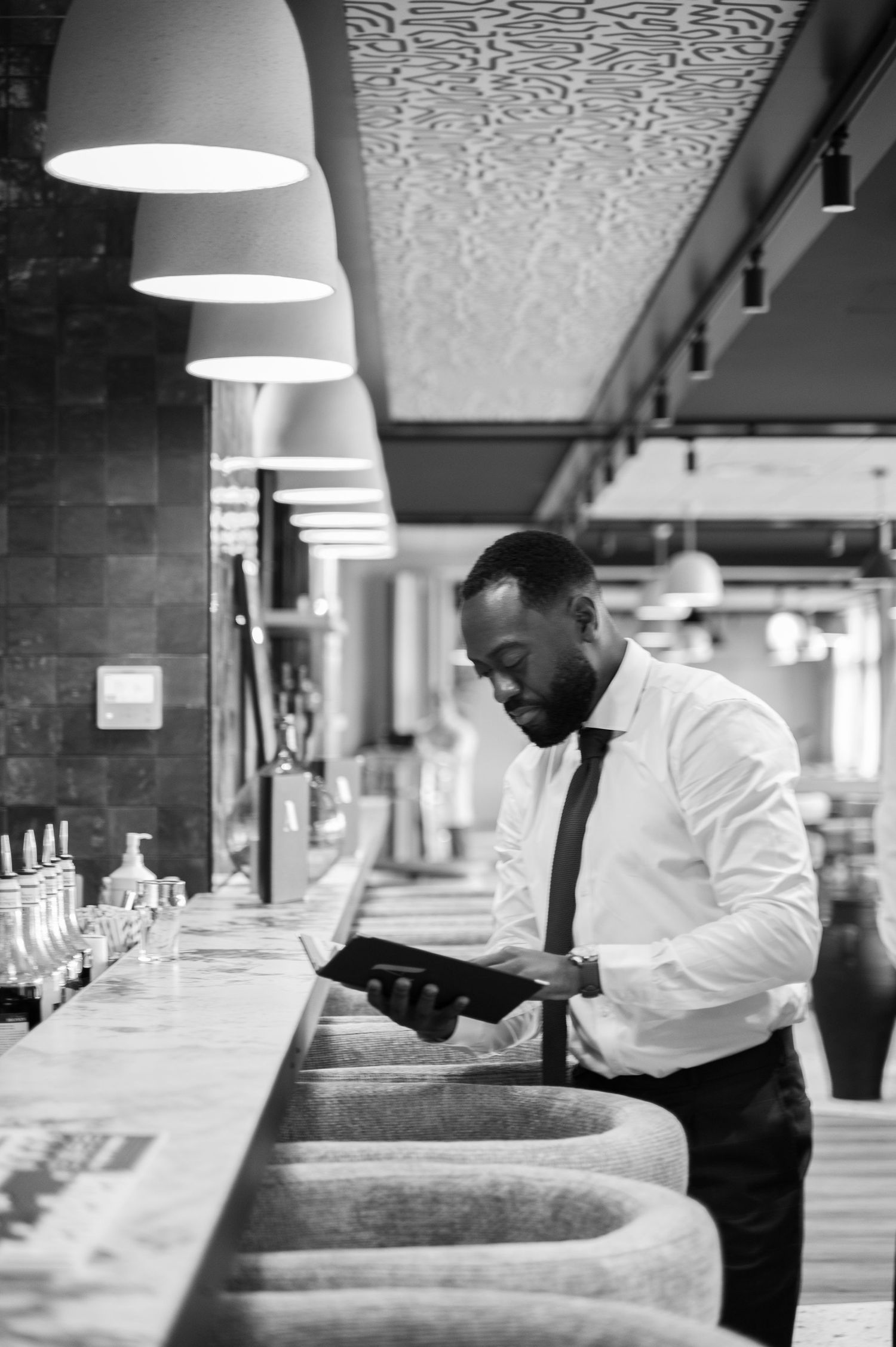 Stylish Black man in white shirt and tie reviews a menu at a modern restaurant bar counter.