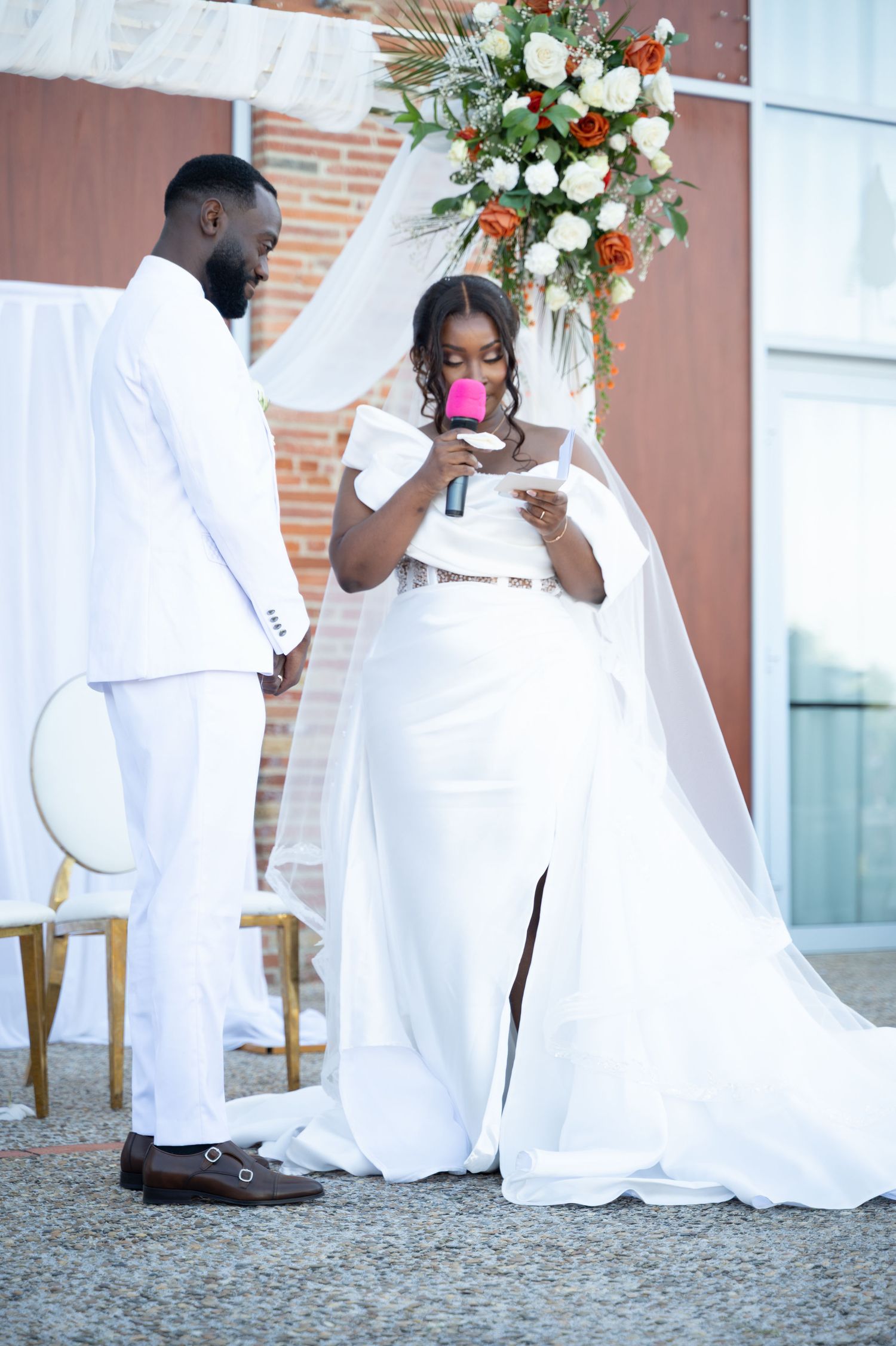 Bride in elegant white gown reads wedding vows while holding pink microphone outdoors.