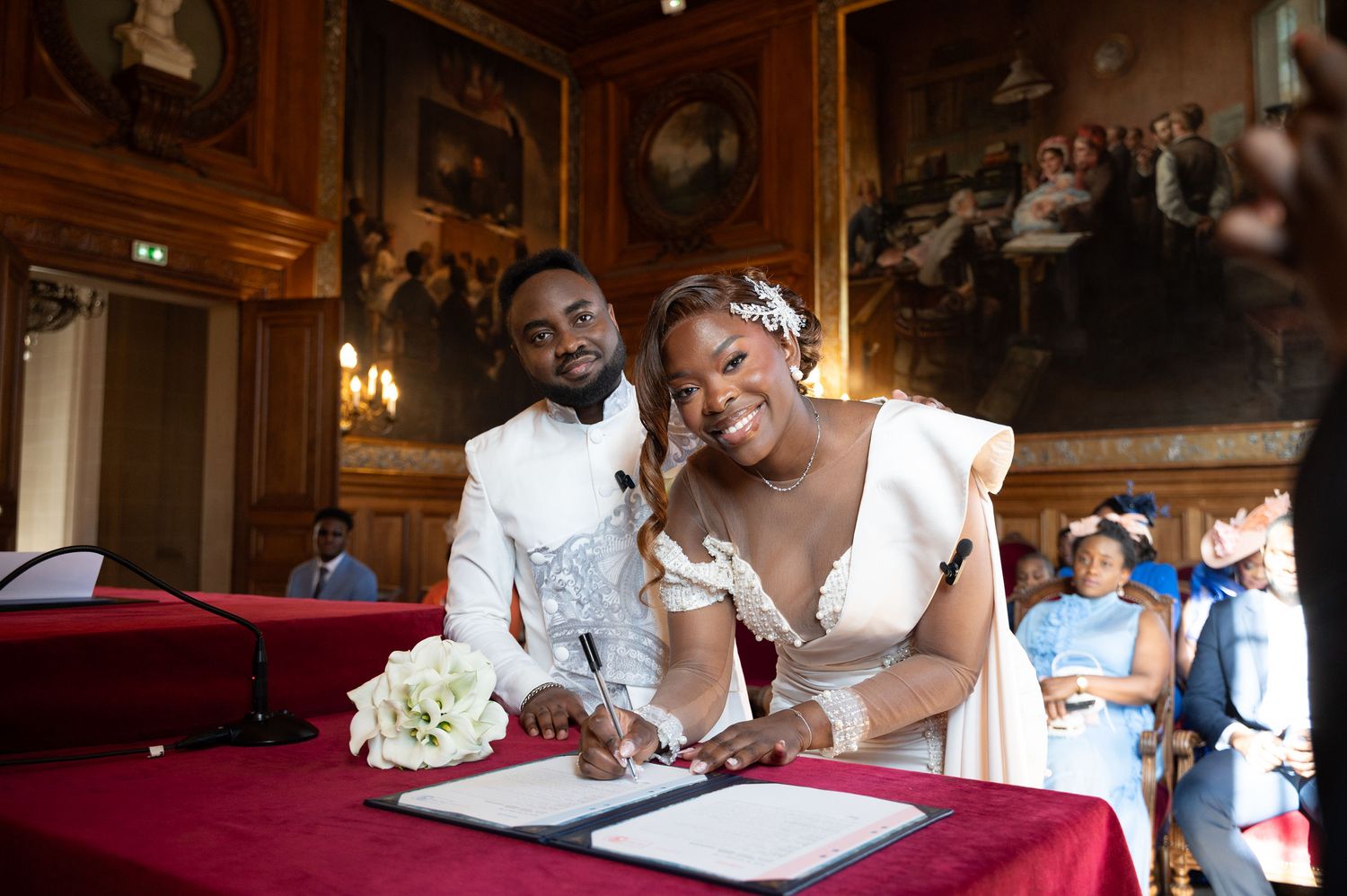 Smiling newlyweds signing their marriage certificate at a ornate table in a grand decorated hall.