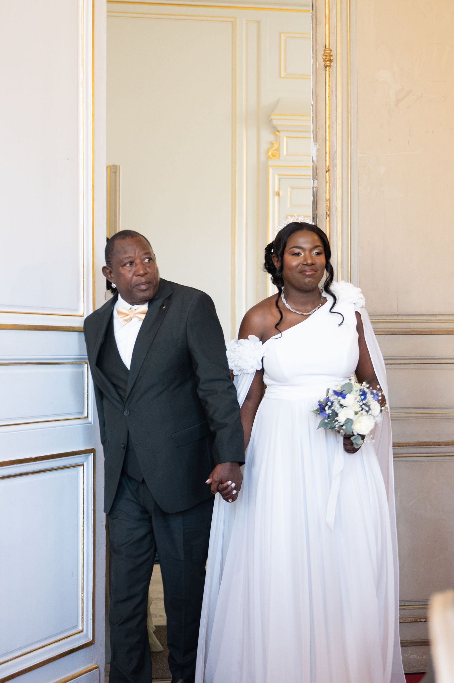 Bride in white one-shoulder gown and groom in black suit holding hands at elegant wedding venue.
