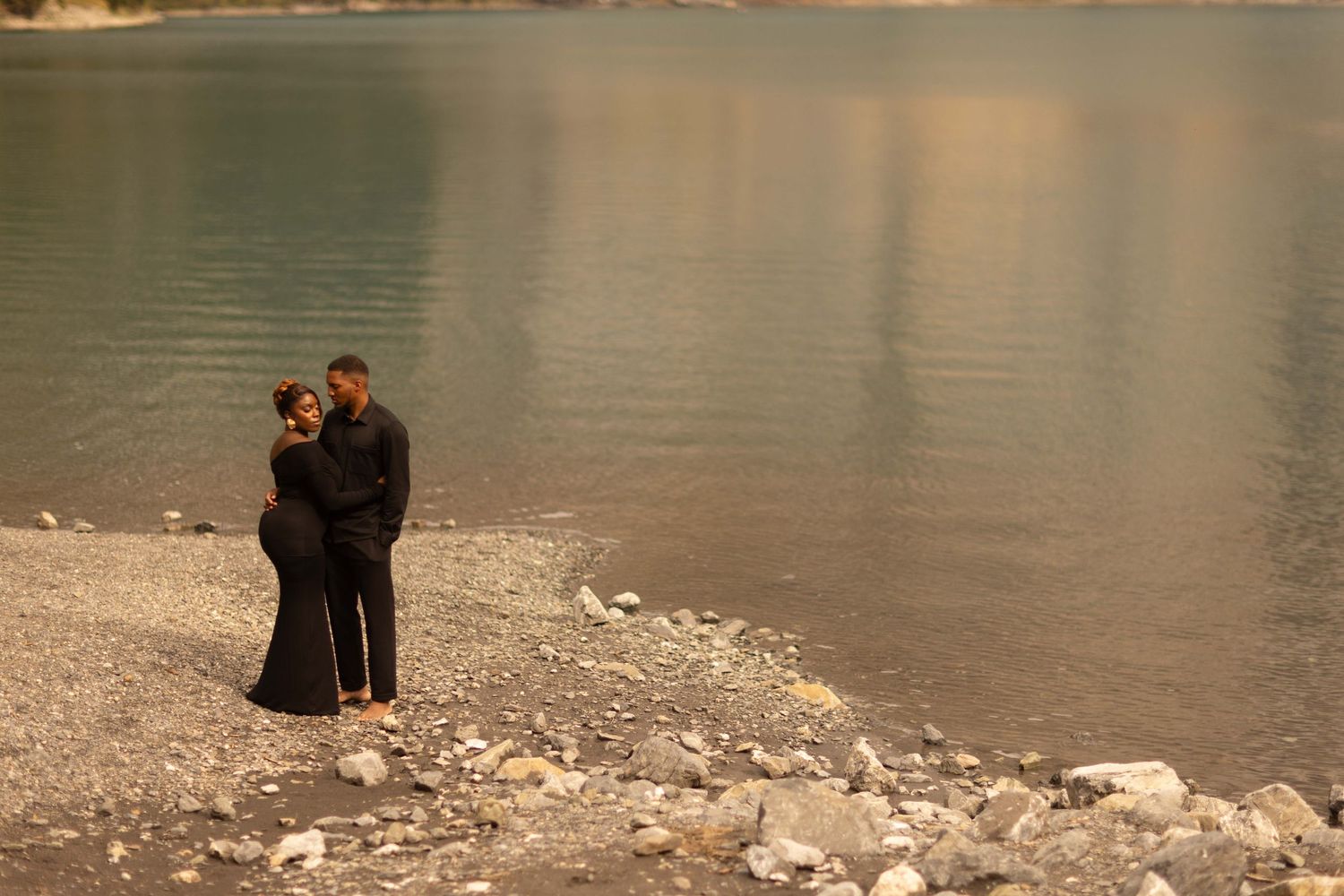 Couple embracing in black outfits on rocky shoreline beside calm turquoise lake waters.