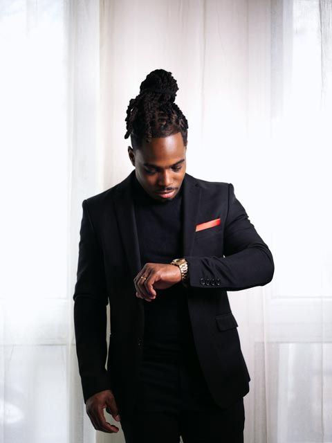 Stylish man in black suit with locs updo checking gold watch, red pocket square accent.