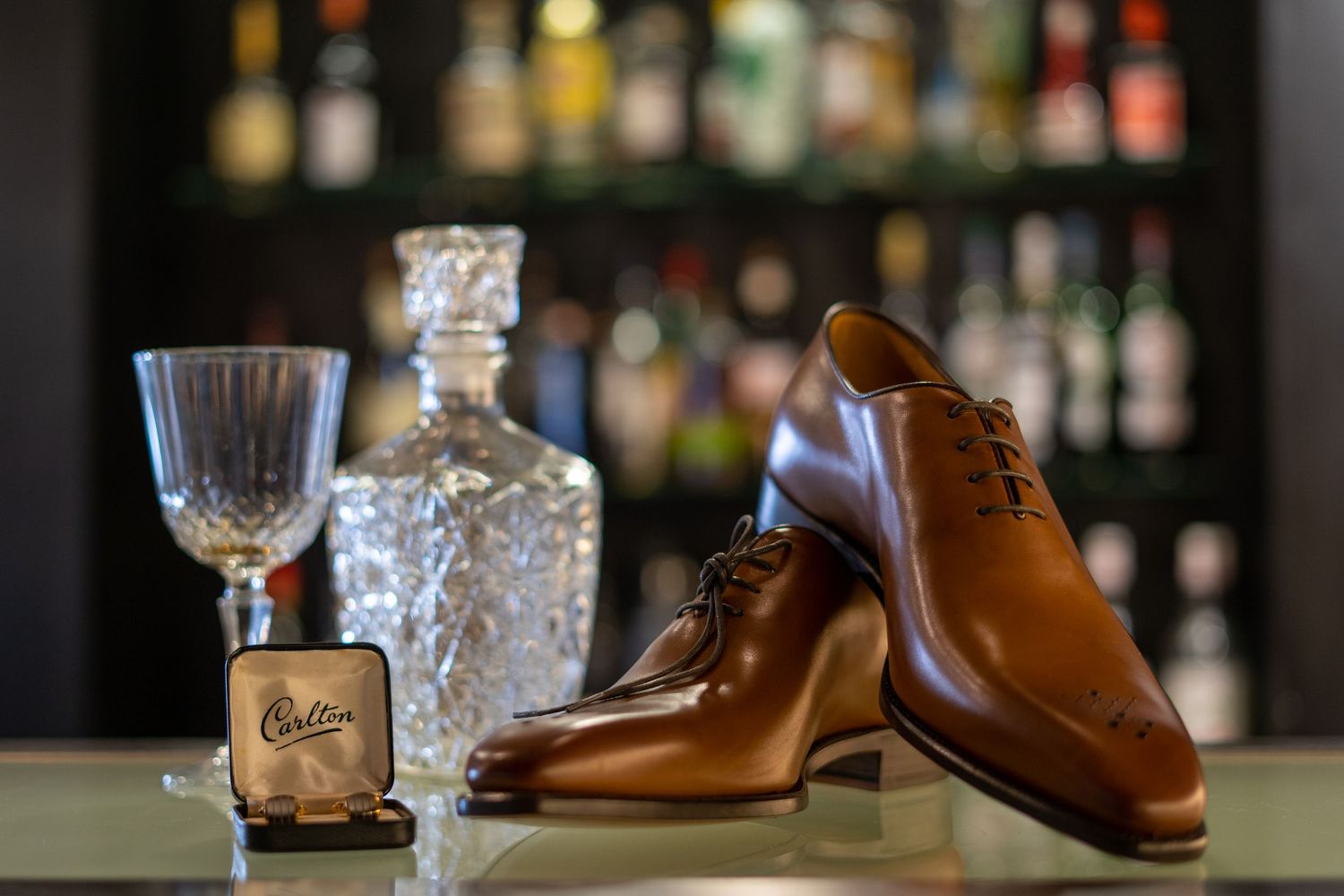 Polished tan Carlton dress shoes displayed beside a crystal decanter and glass on a reflective surface.