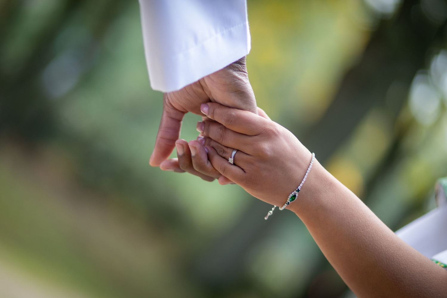 Close-up of couple holding hands showing diamond engagement ring and silver bracelet with green bokeh background.