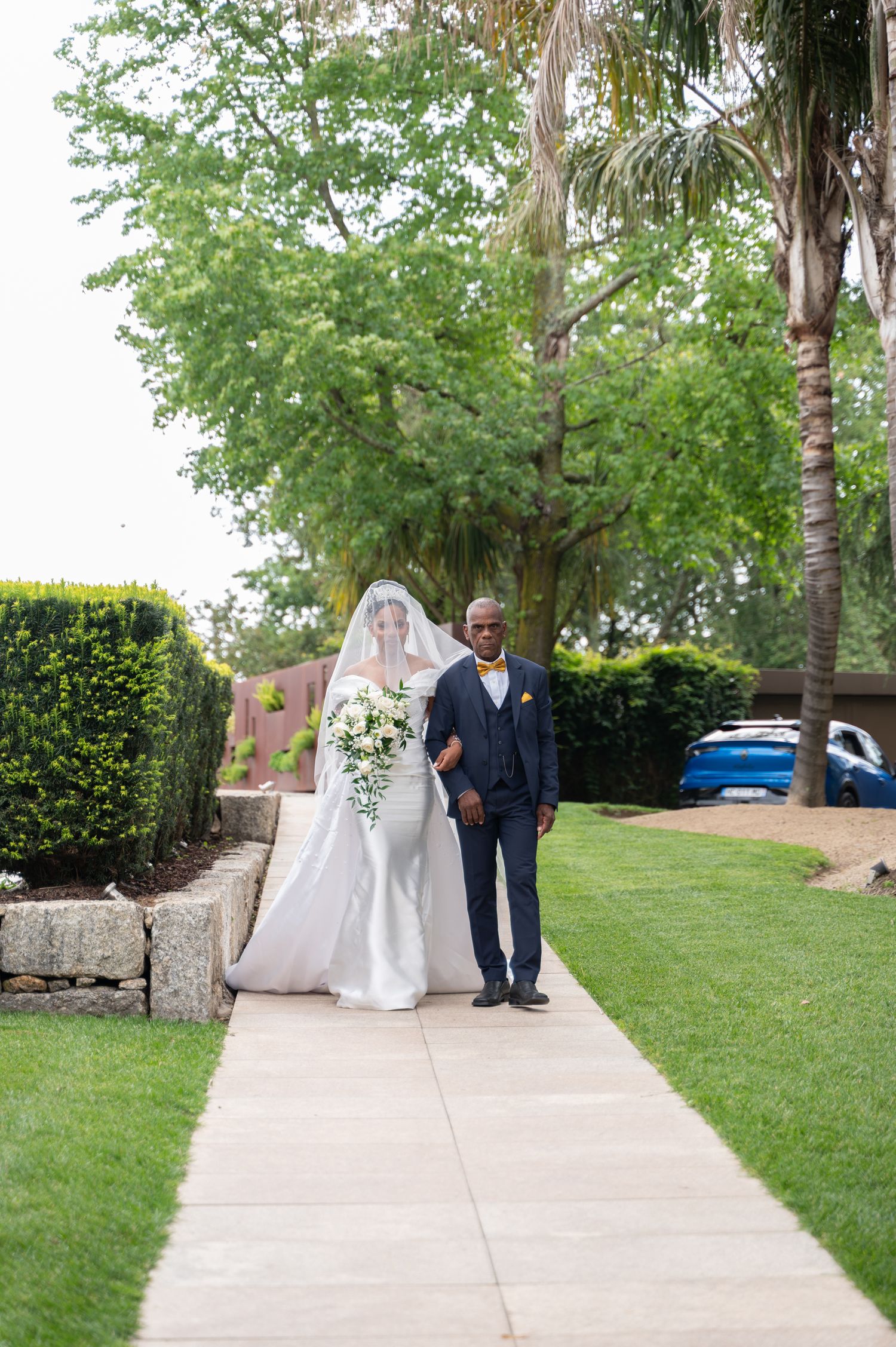 Bride in white gown and veil walks along a garden path escorted by her father on her wedding day.
