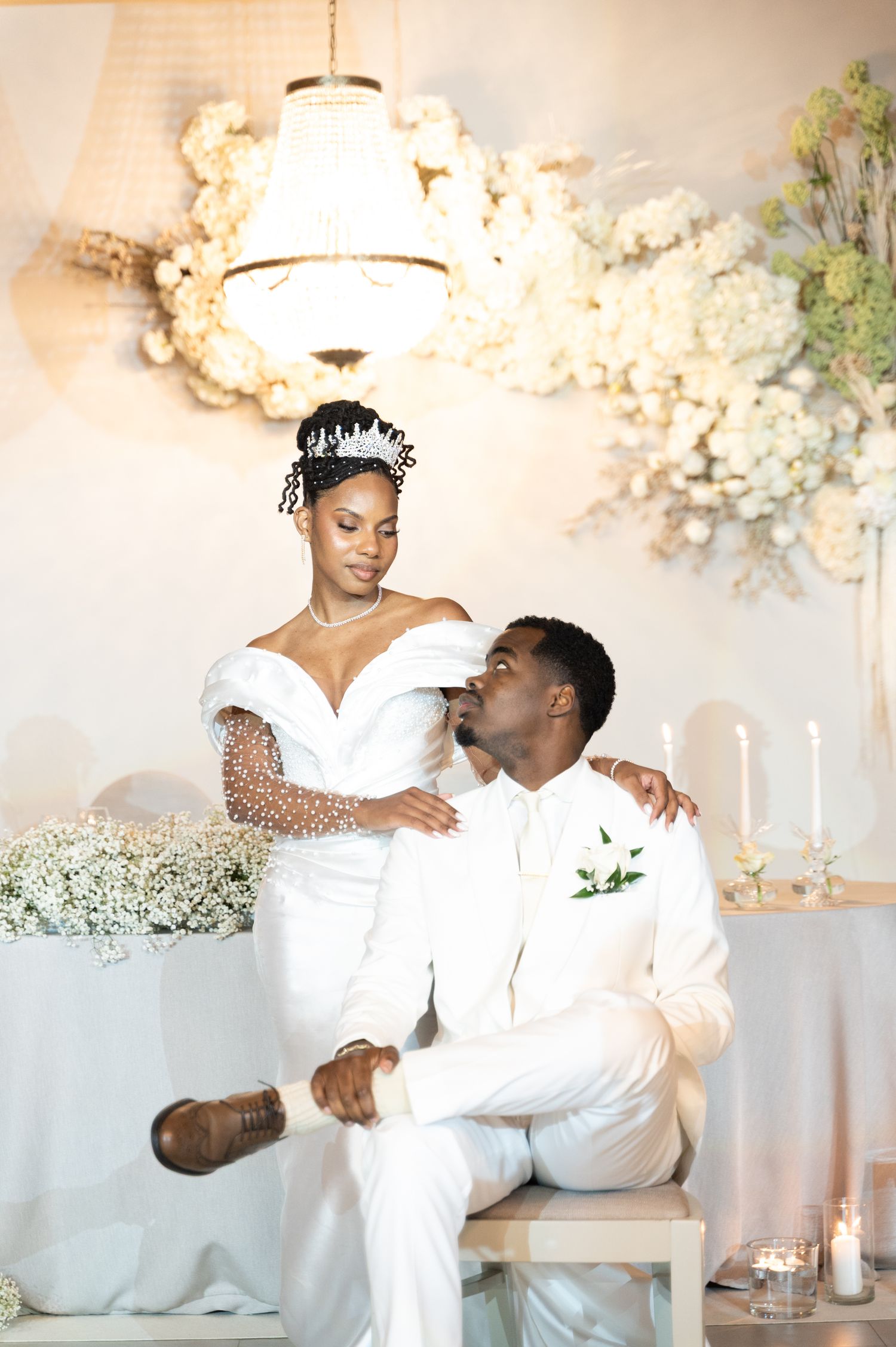 Bride in pearl-embellished gown and crown sits beside groom in white suit beneath a floral chandelier backdrop.