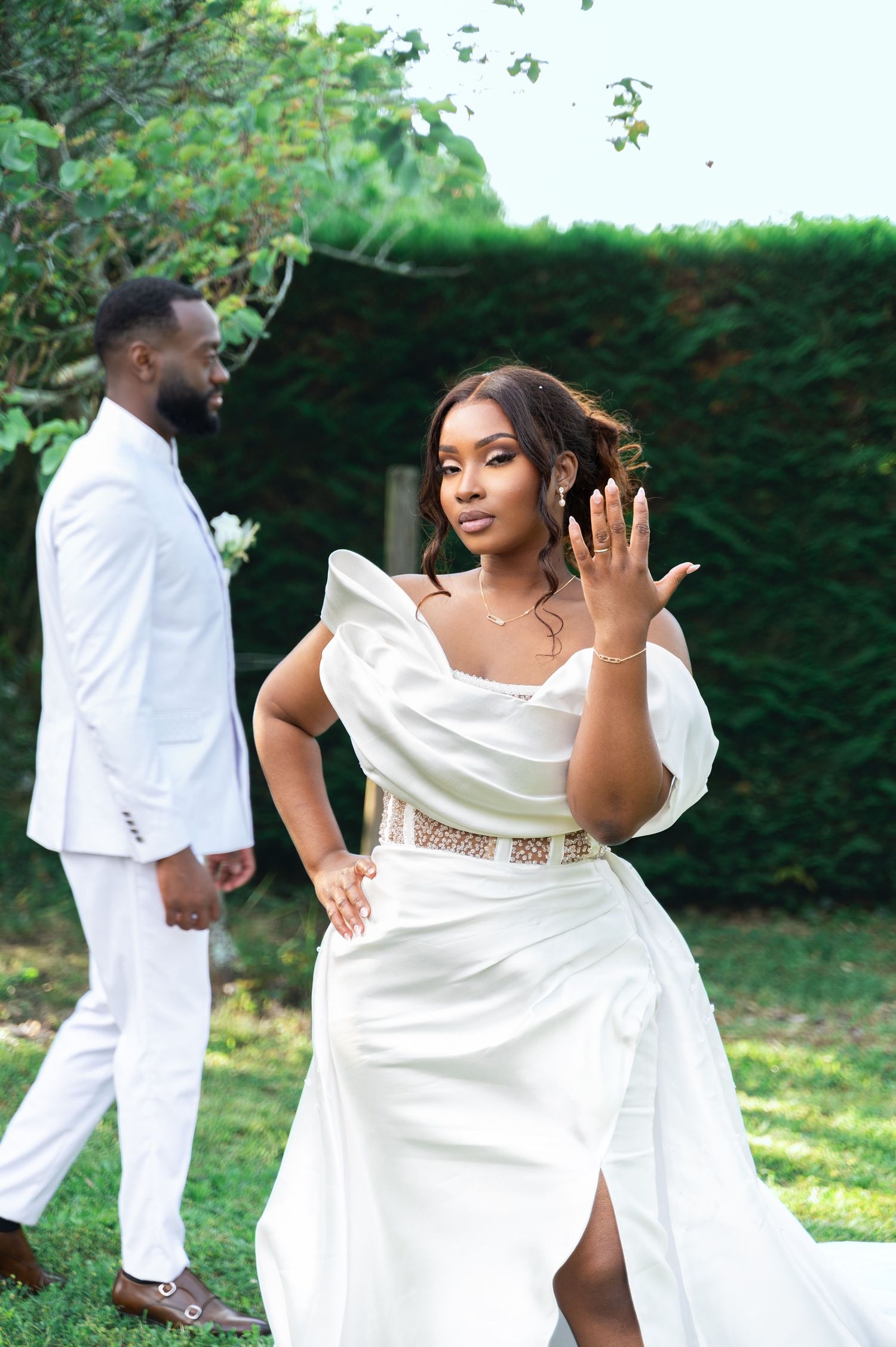 A radiant Black bride in a white off-shoulder gown shows her ring while her groom stands behind her.