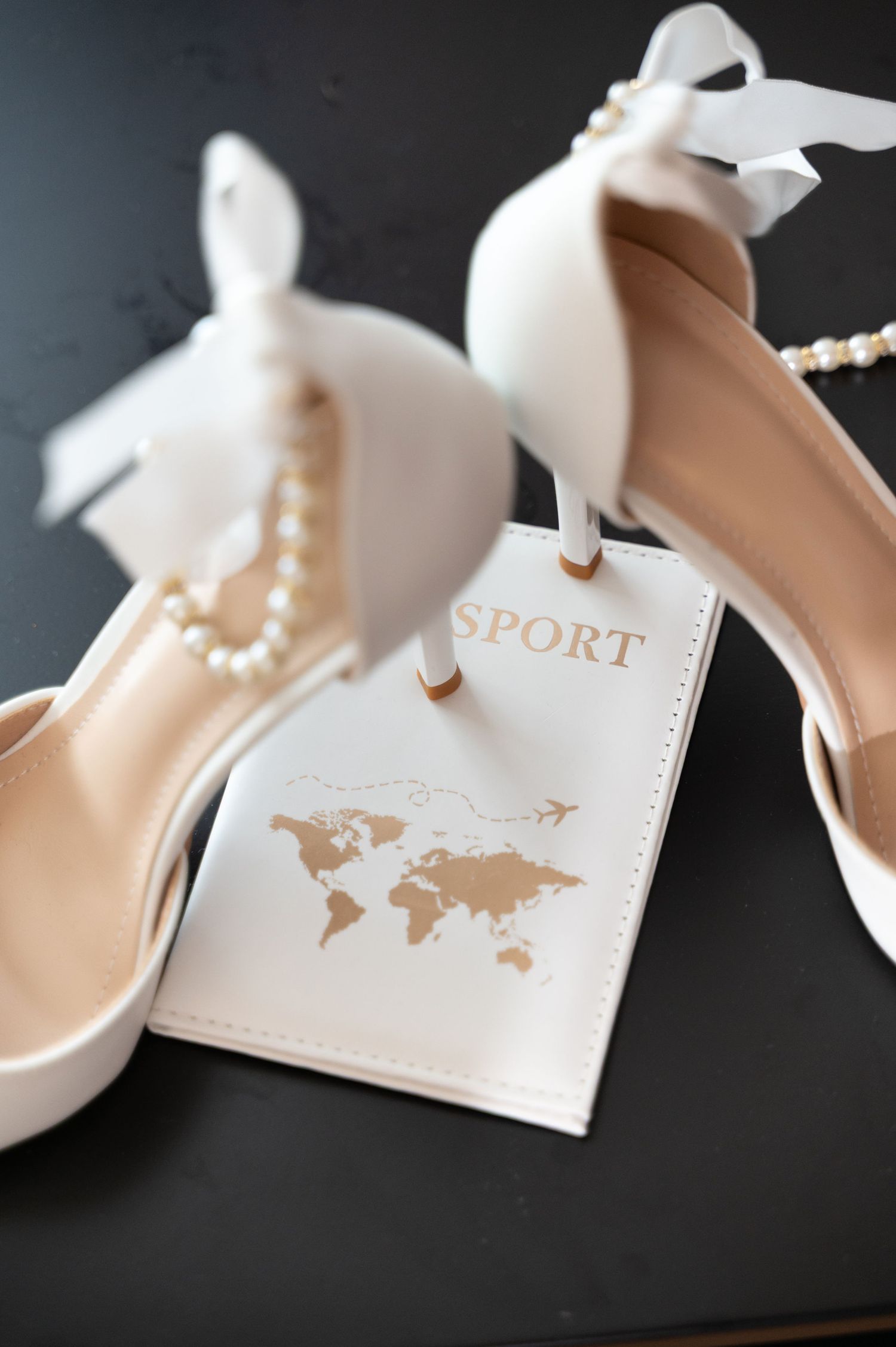 White bridal heels with pearl necklace and passport holder on dark surface.