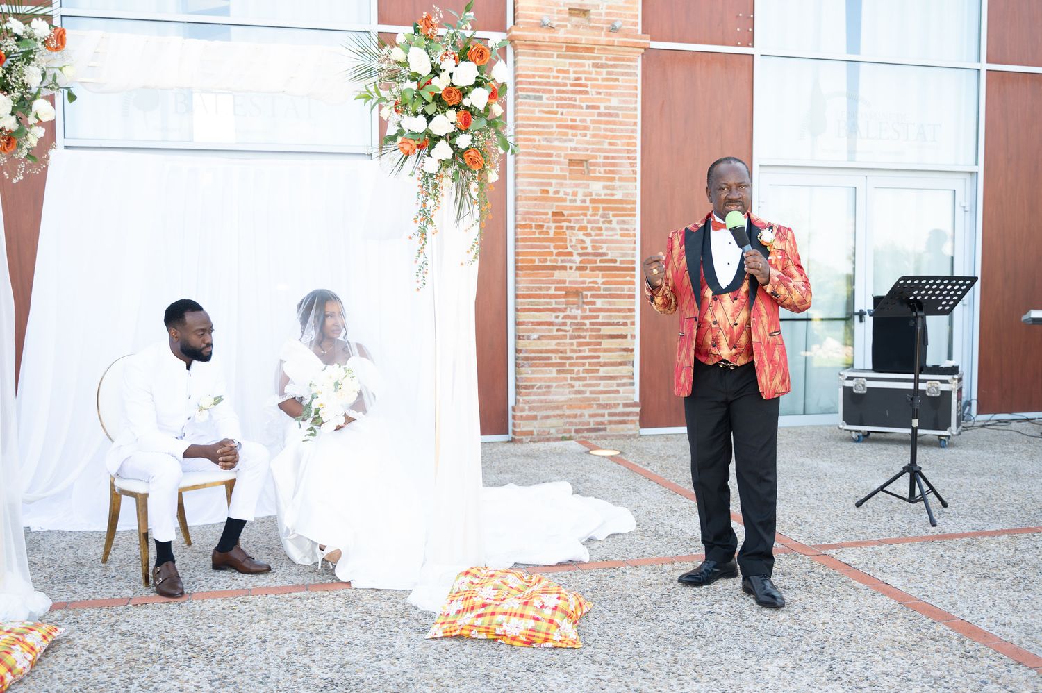 Wedding officiant in red jacket speaks to seated bride and groom at outdoor ceremony.