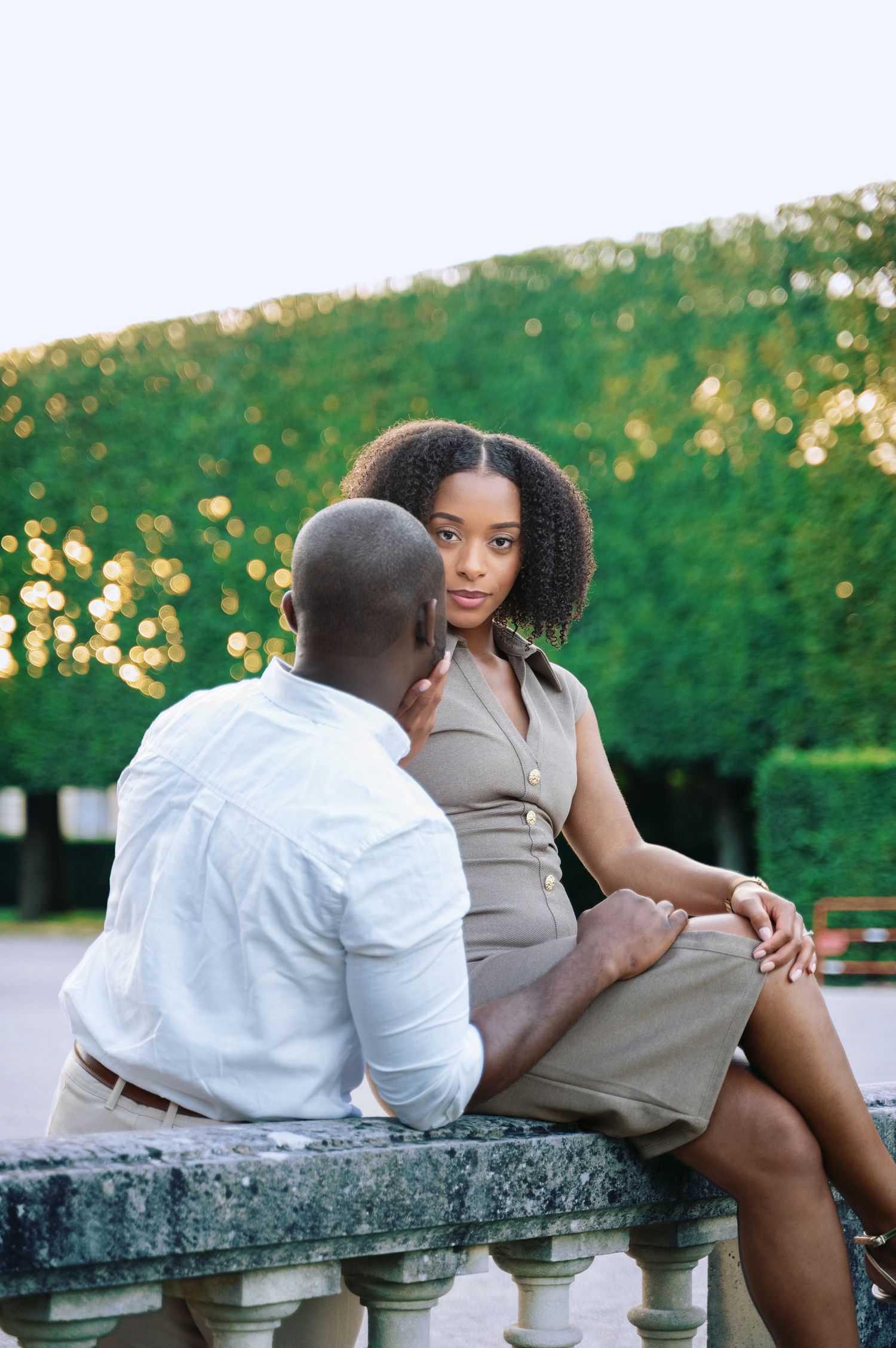 A Black couple shares an intimate moment seated on a stone balustrade in a sunlit formal garden.
