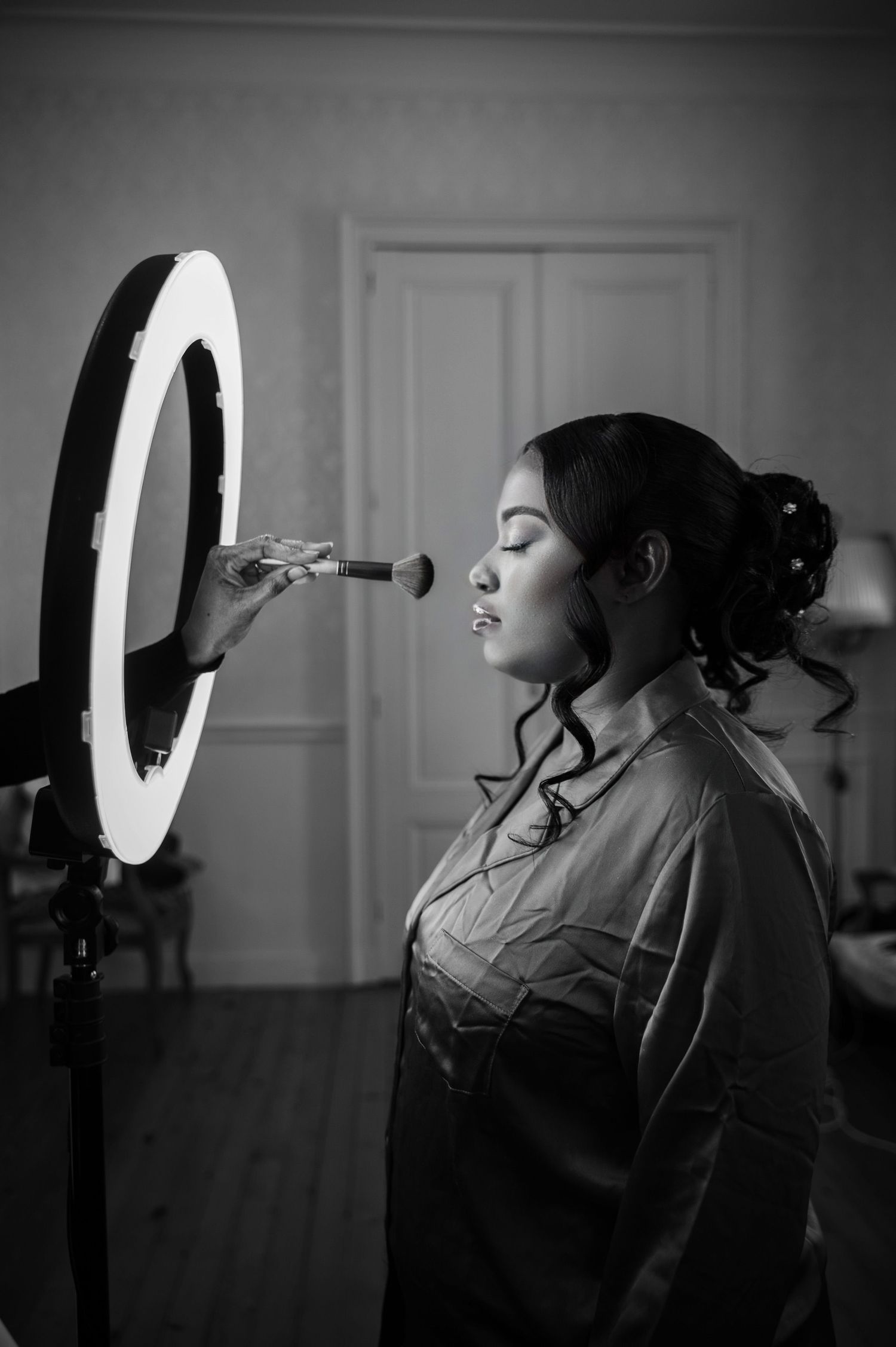 A Black bride gets makeup applied with a brush under a ring light in a black and white portrait.