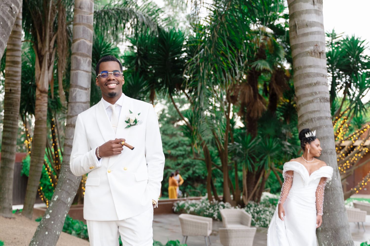 Groom in white suit points finger while bride in white gown stands behind near palm trees.