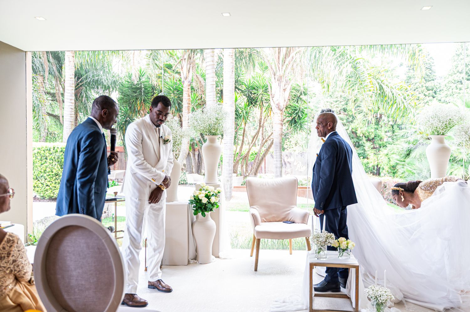 Groom in white suit stands at altar with officiant and guests during an elegant indoor wedding ceremony.
