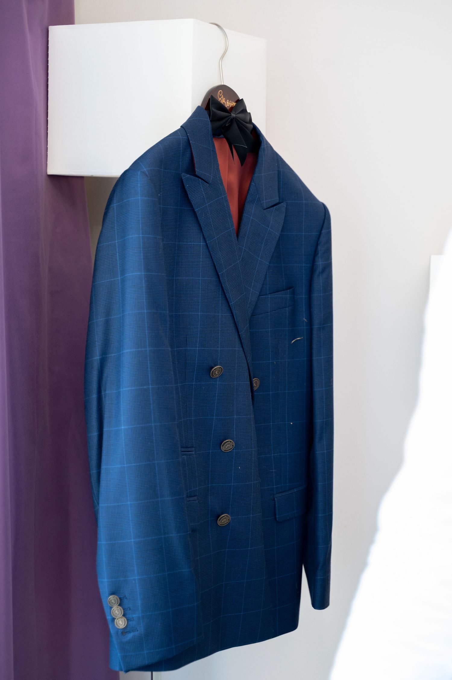 Double-breasted navy blue plaid suit jacket hanging on wooden hanger against white wall for groom.