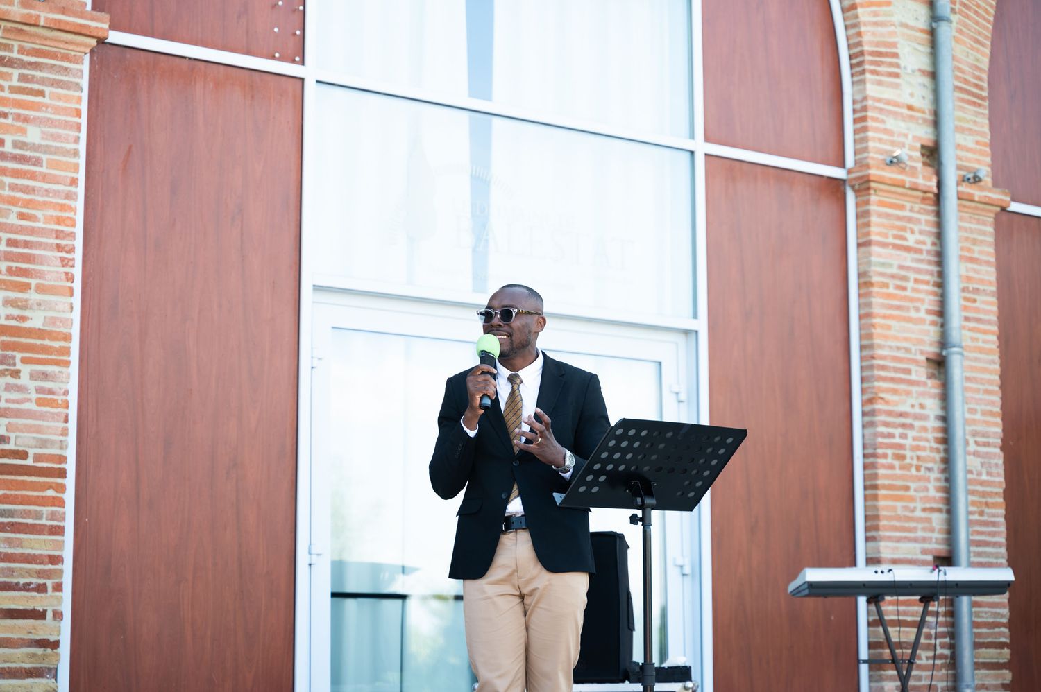 Man in blazer speaks at outdoor wedding ceremony podium with music stand.