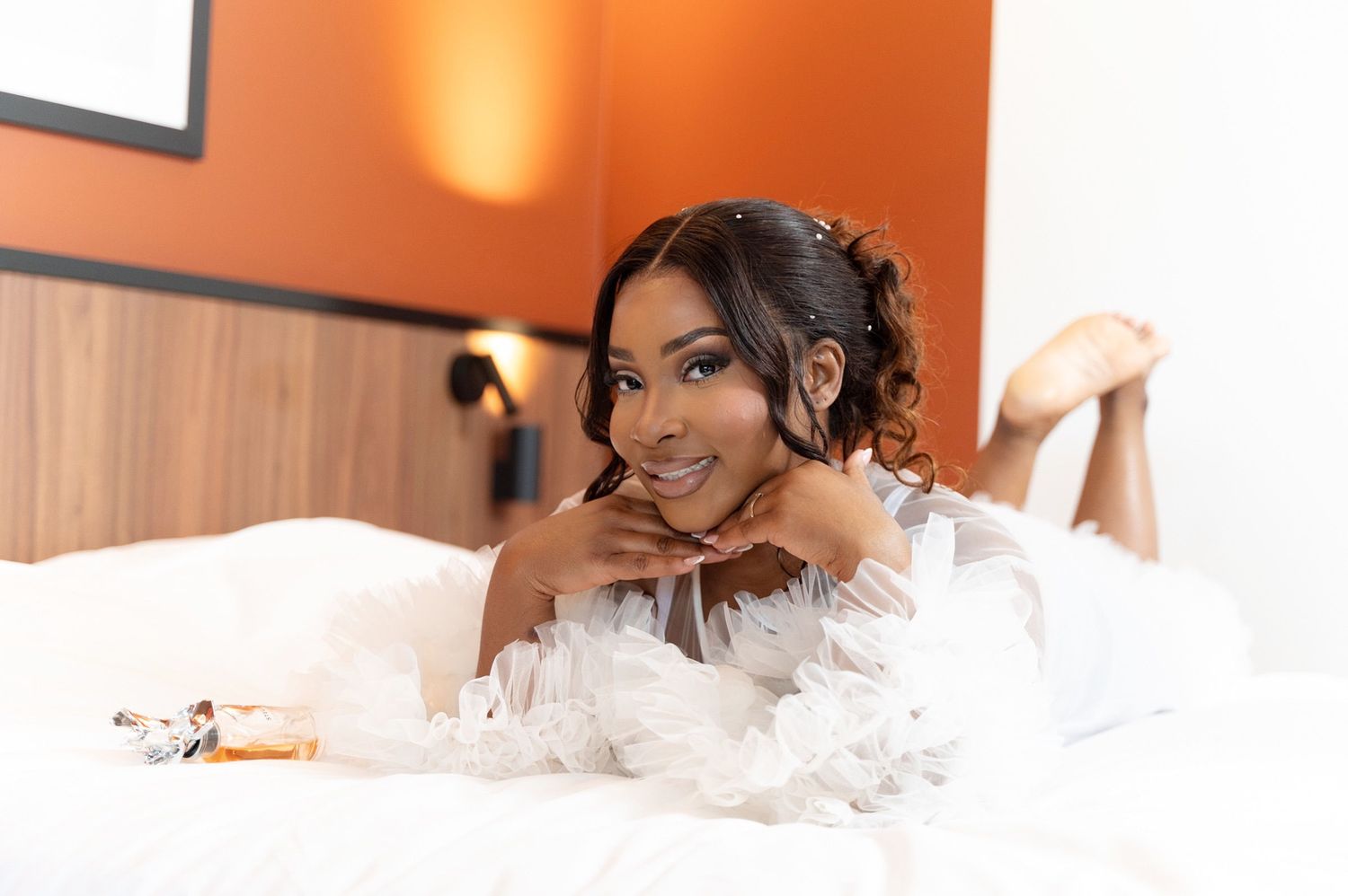 Radiant bride in white feathered robe poses on a white bed with a perfume bottle nearby.