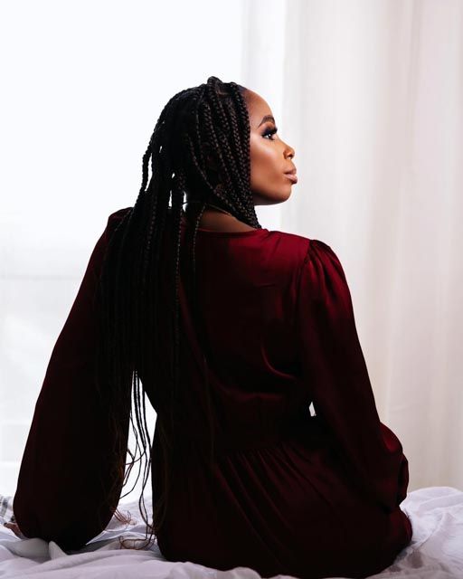 Woman in burgundy dress with long box braids sits gracefully on bed, gazing toward window light.