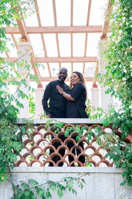 Couple in black outfits smiling on a pergola balcony surrounded by lush green vines and decorative lattice.