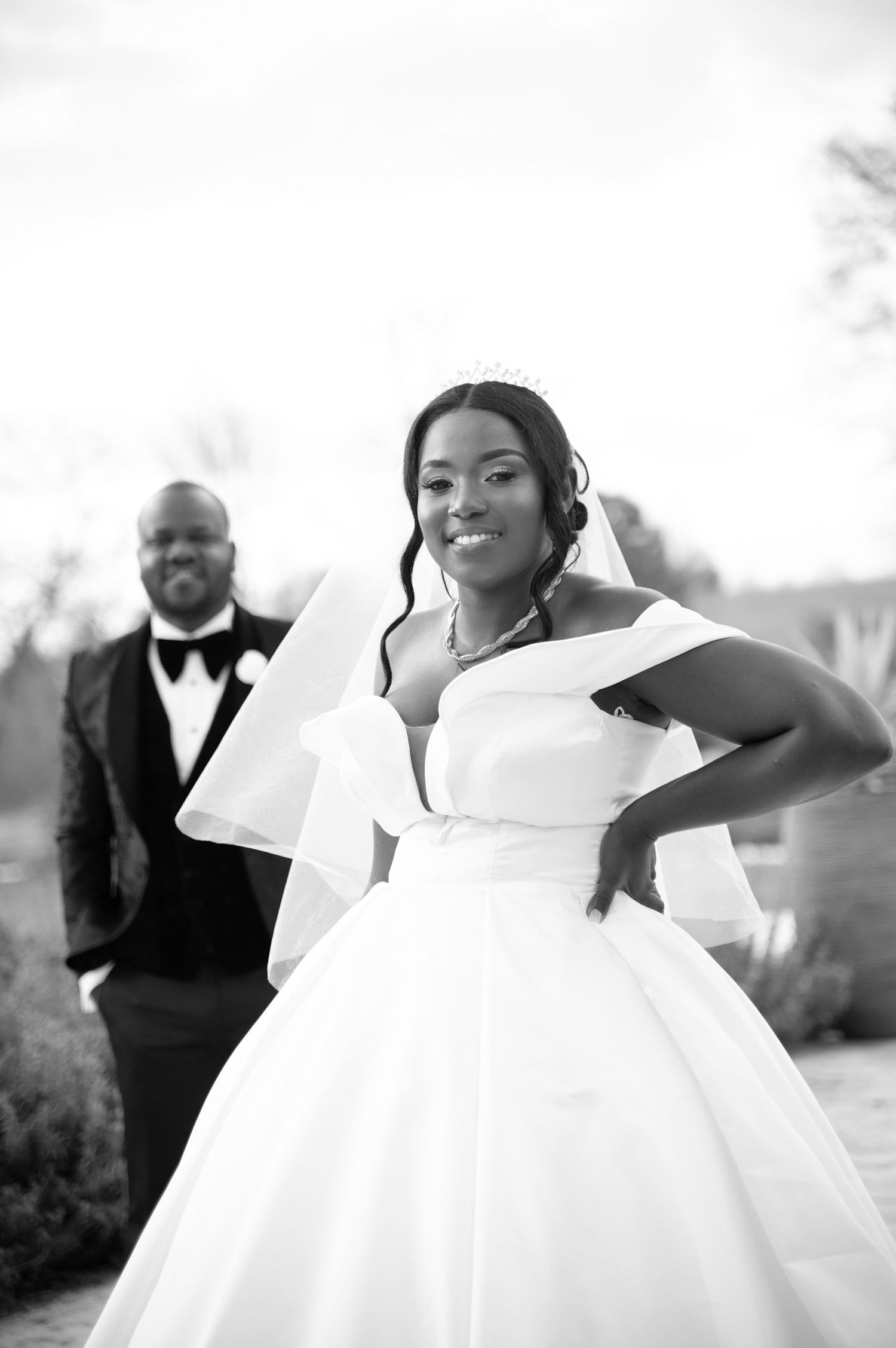 Black and white photo of smiling bride in tiara and ballgown posing outdoors with groom in background.