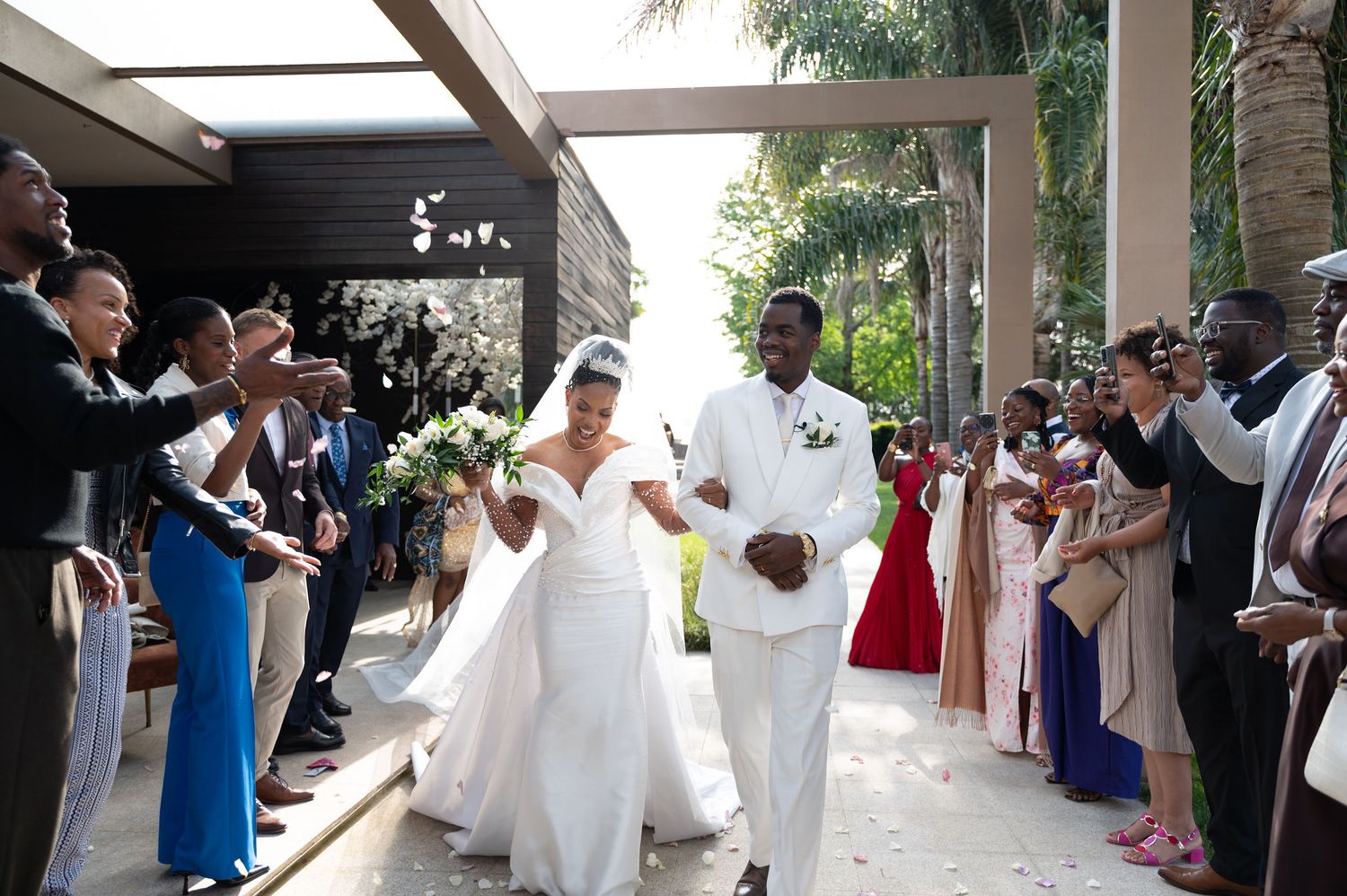 Joyful newlyweds walking down the aisle surrounded by cheering guests, bride in white gown and groom in white suit.