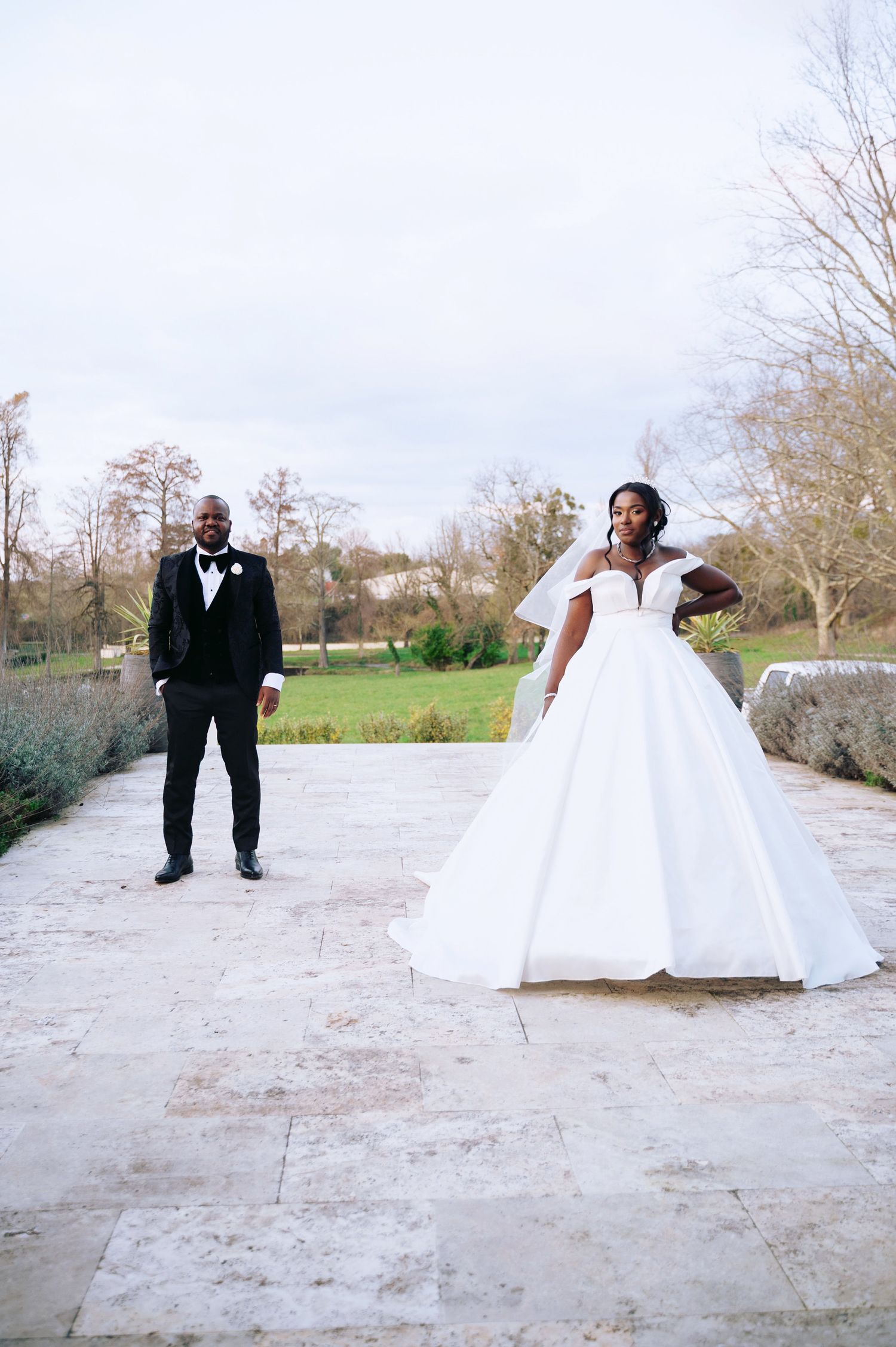 Bride in voluminous off-shoulder ball gown and groom in black tuxedo posing on stone terrace outdoors.
