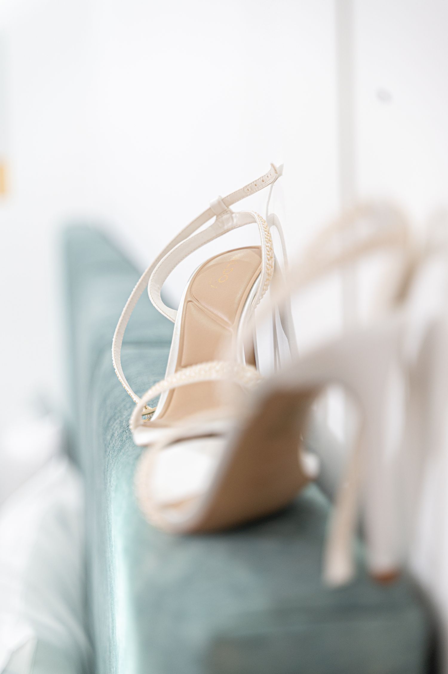 Elegant white strappy bridal heels displayed on a light blue velvet chair.