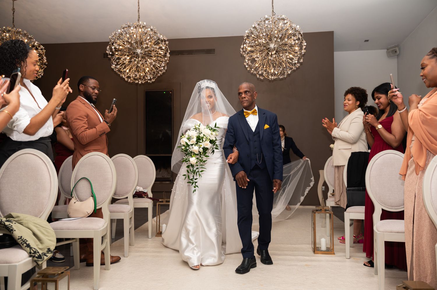 Bride in cathedral veil carrying white bouquet walks the aisle with escort under crystal chandeliers.
