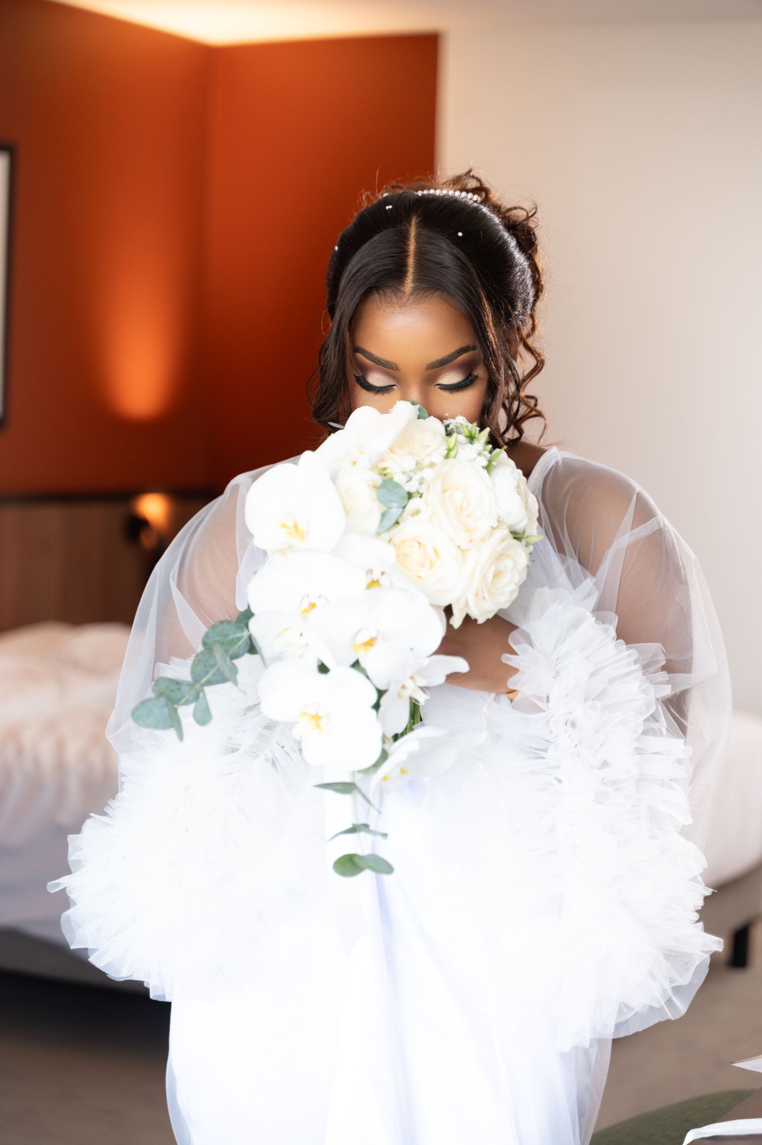 Black bride in white tulle gown smells large white rose and orchid bouquet in hotel room setting.