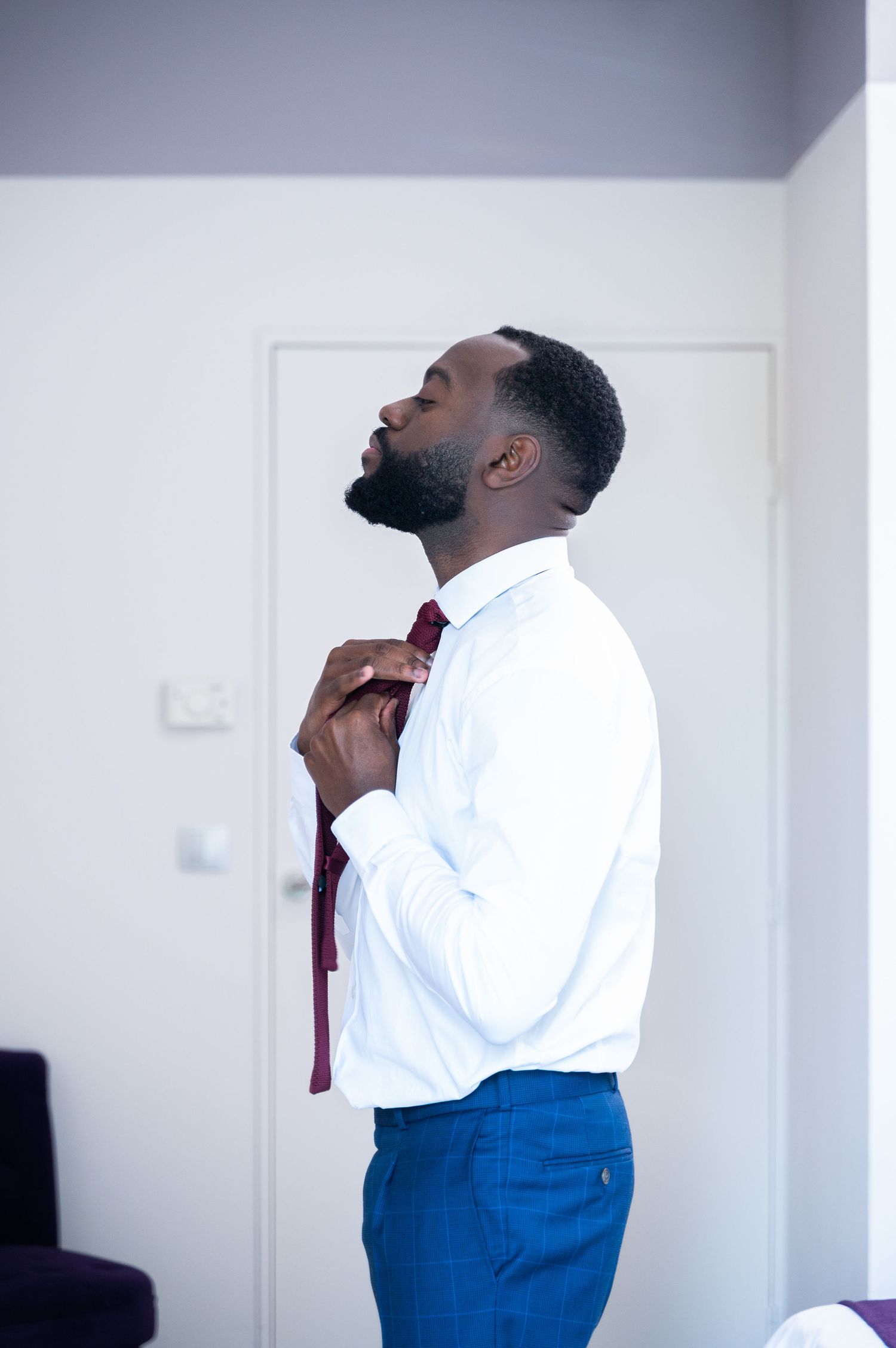 Handsome groom in white dress shirt and blue pants adjusting his burgundy tie before wedding ceremony.
