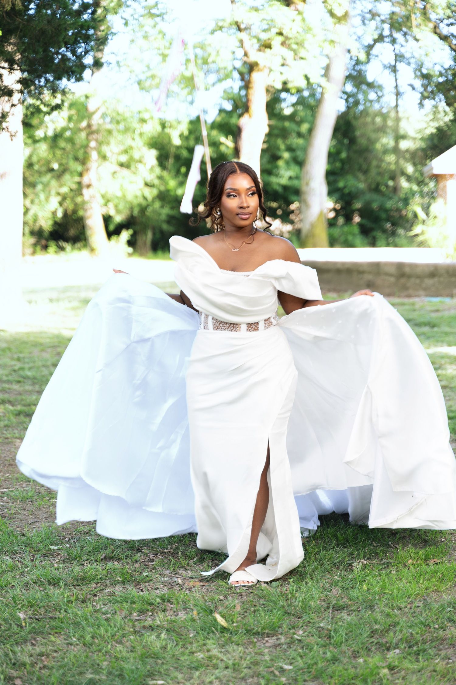 Beautiful bride in white off-shoulder wedding dress with slit and gold belt spreads train on green lawn.