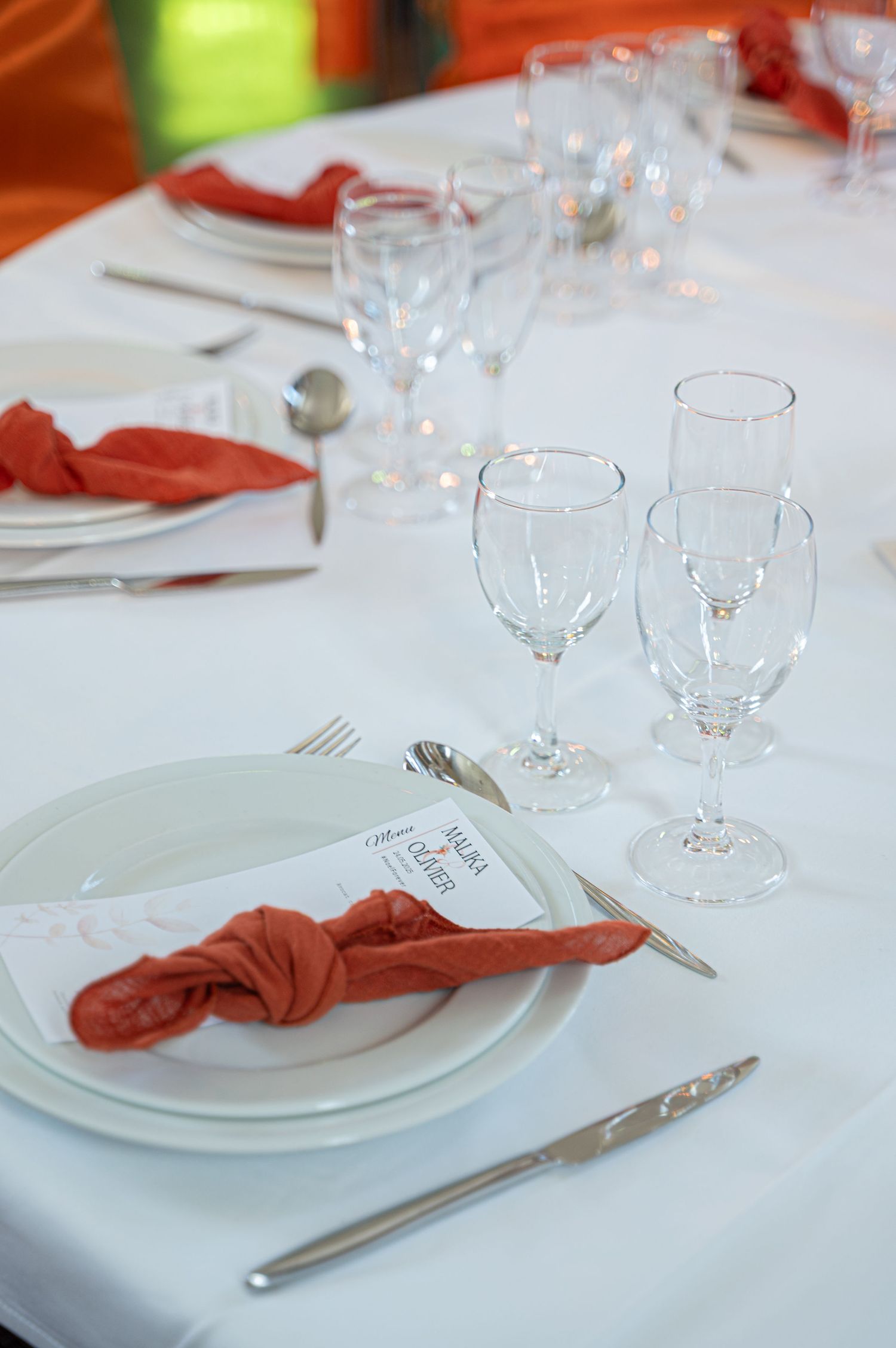 Elegant wedding table setting with white plates, orange napkins, silverware, and crystal wine glasses.