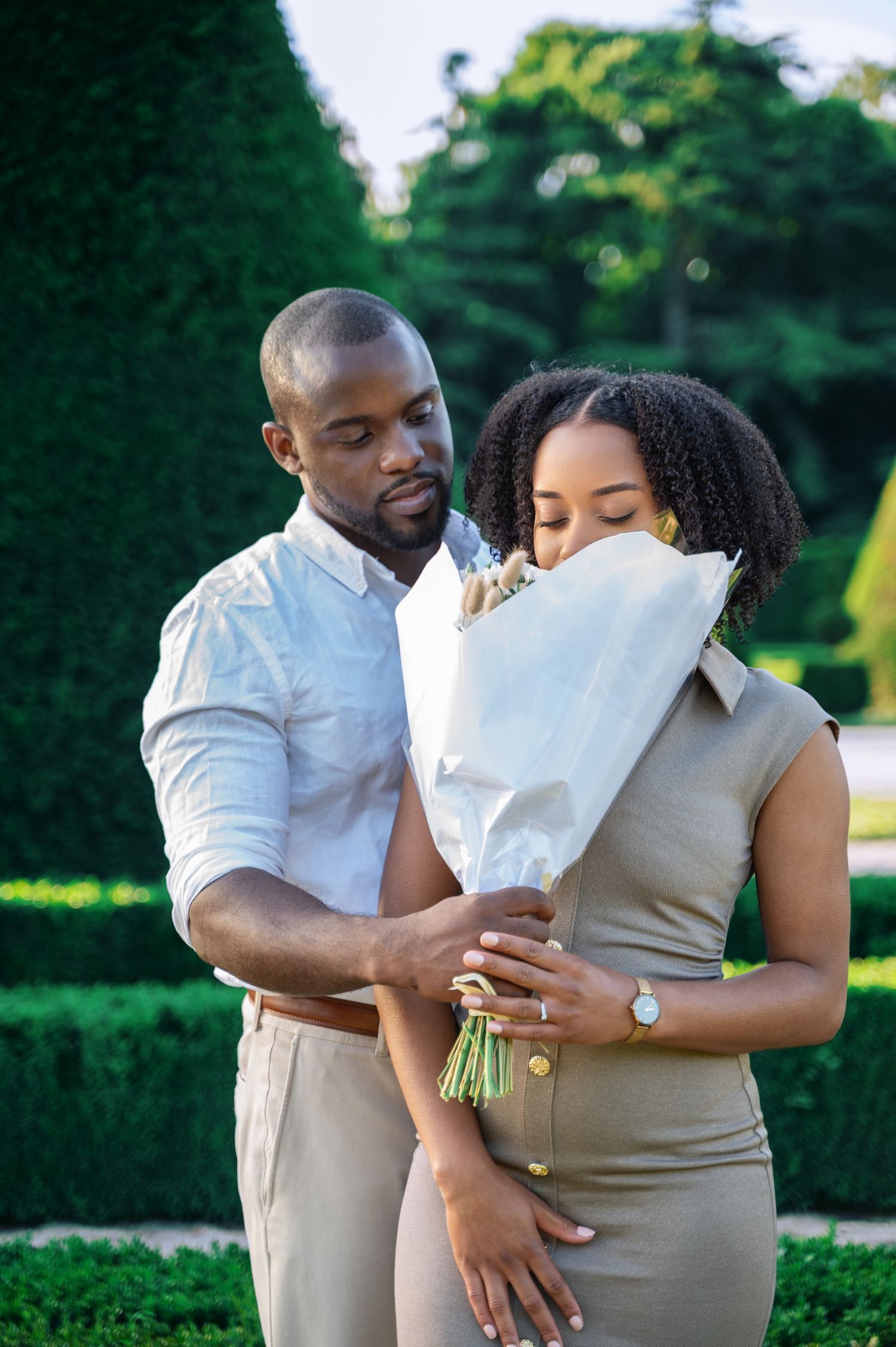 Man presenting white-wrapped flower bouquet to woman in formal garden setting.