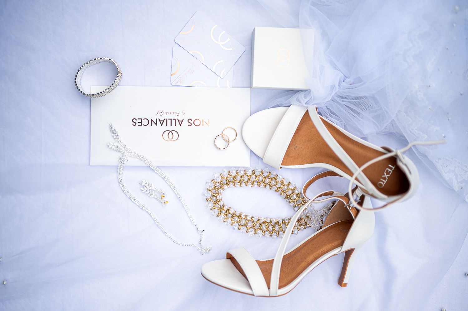 Bridal flat lay featuring white heeled sandals, pearl bracelet, rings, and wedding stationery on veil.