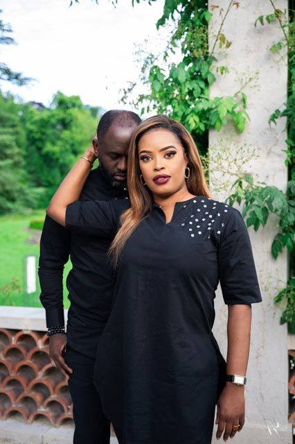 Stylish couple in matching black outfits posing outdoors with lush green foliage background.