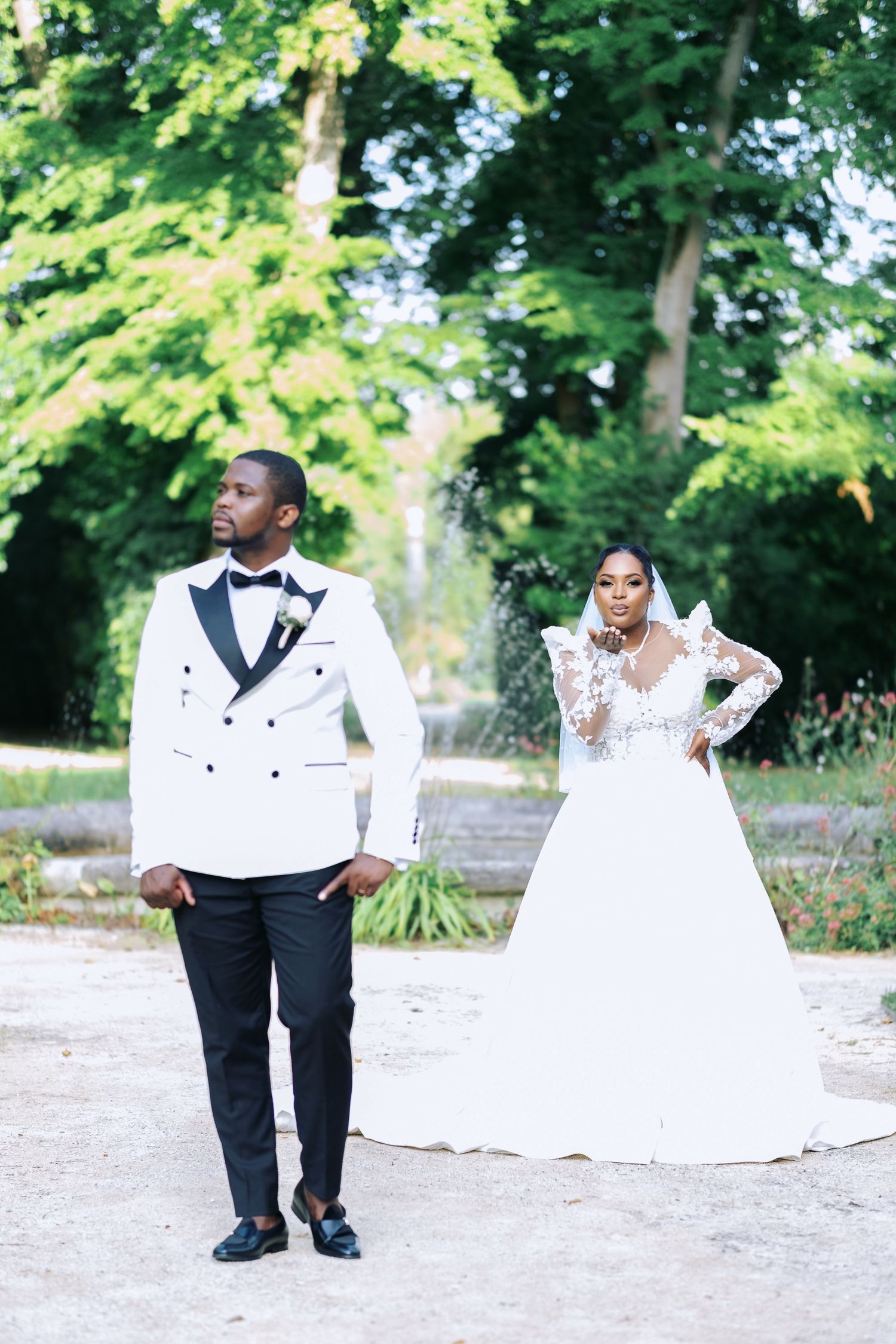 Black couple in elegant wedding attire pose outdoors, groom in white tuxedo, bride in lace gown with long sleeves.